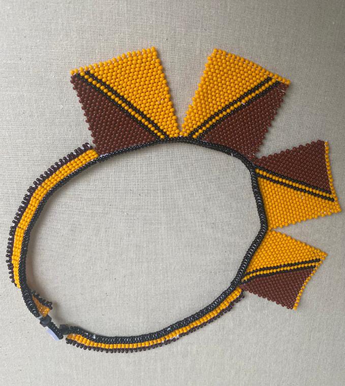 A beaded necklace with triangular fringe pieces in yellow and brown. The striking design, reminiscent of the Weight of Silence collection by Contra.Joburg, features alternating triangles on a neutral fabric background for a bold statement look.