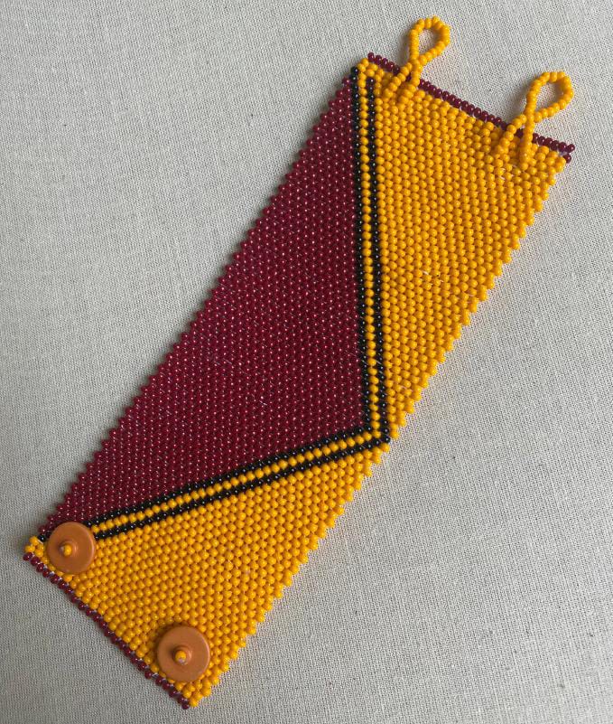 A beaded bracelet with a geometric pattern: maroon, yellow, and black beads form angled shapes. Inspired by Contra.Joburg, it features two orange buttons for fastening and yellow beaded loops, resting on a light fabric background.