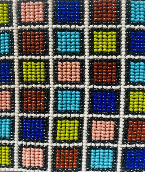 A close-up of a colourful beaded pattern featuring a grid of small squares. Reminiscent of Contra.Joburg’s vibrant style, each square is filled with blue, yellow, pink, brown, or orange beads bordered by black and white in a geometric mosaic.