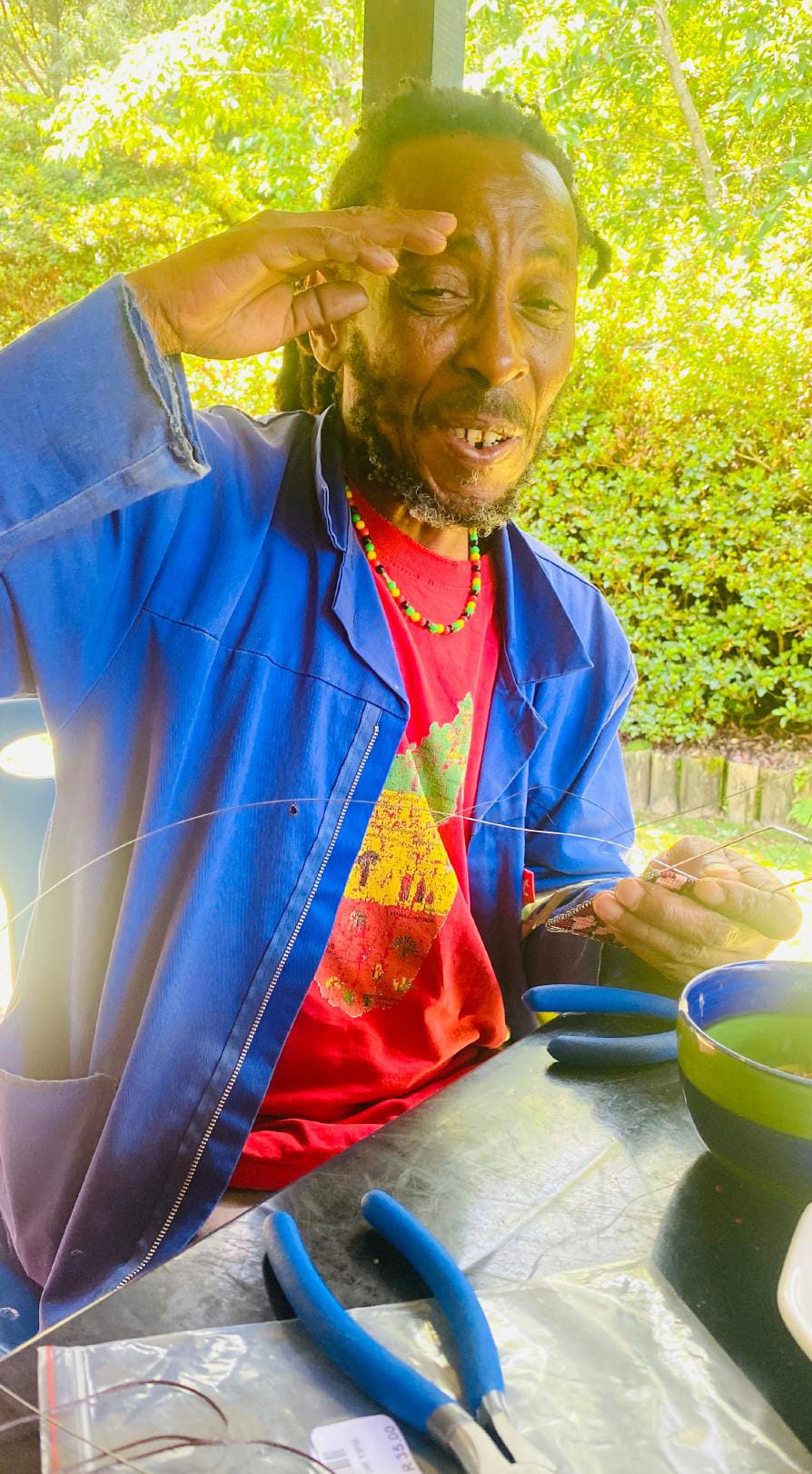 A man with dreadlocks, wearing a blue jacket over a red shirt with a colourful print, sits outdoors by a table. He works intently on wire art or jewellery—perhaps inspired by the Weight of Silence—amid lush greenery and bright light.