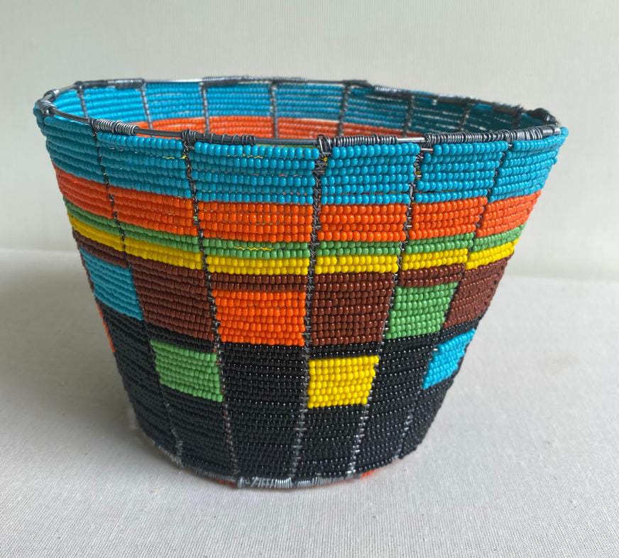 A colourful, handwoven basket from Contra.Joburg features small, square glass beads arranged in a chequerboard pattern with blue, orange, yellow, green, black, and brown sections. The wide opening and tapered sides evoke the delicate Weight of Silence.