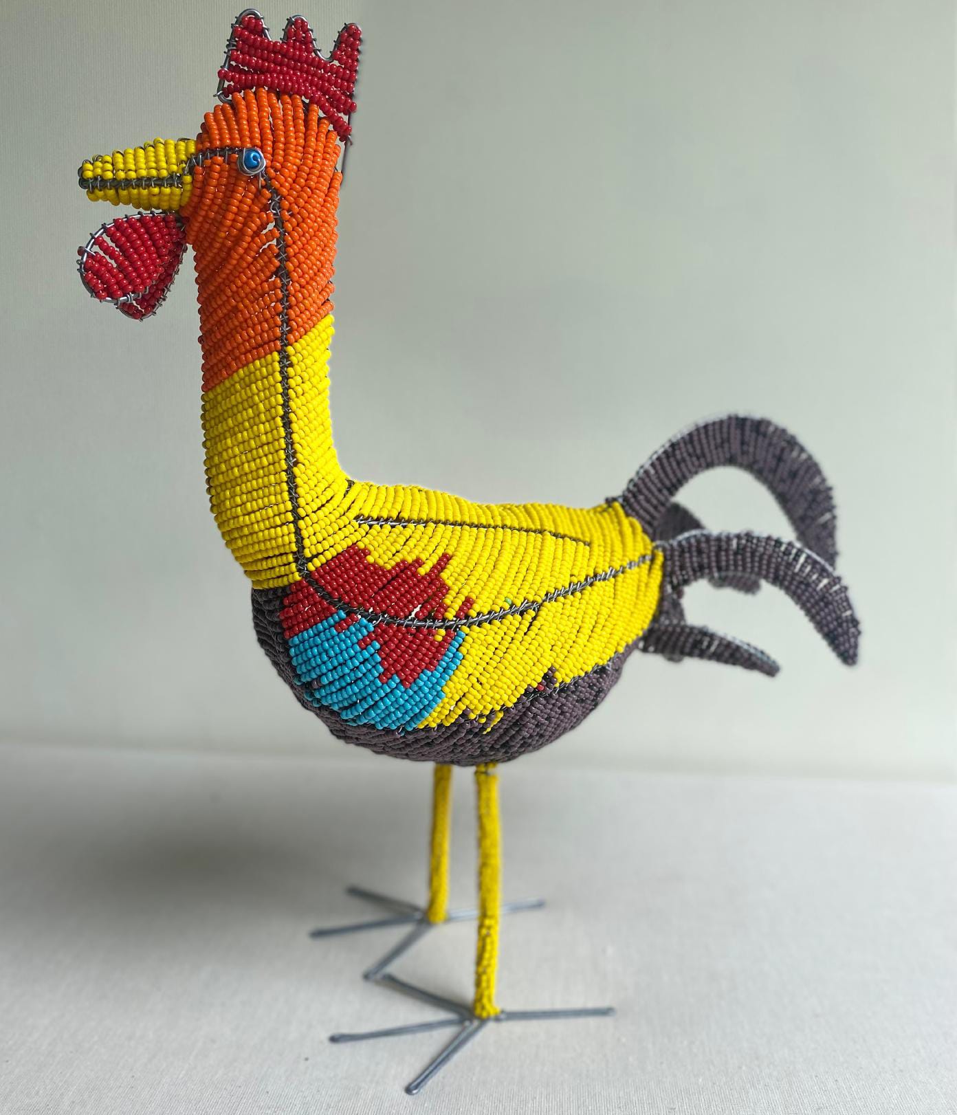 A colourful cockerel sculpture from Contra.Joburg, made of small, shiny beads in yellow, orange, red, blue, and grey, stands upright with a red comb and wattles—its vibrant presence breaks the Weight of Silence against a plain light background.