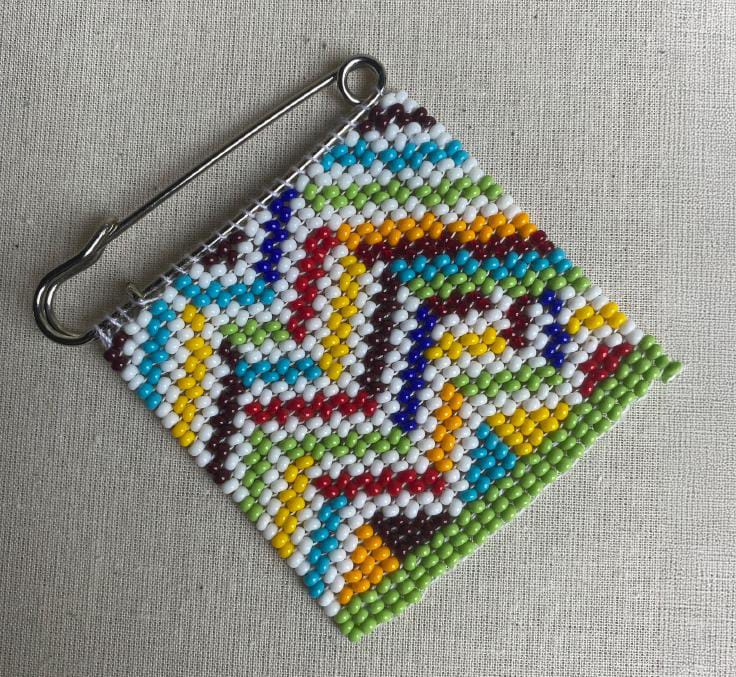 A square beaded patch attached to a large safety pin, featuring zigzag patterns in vibrant hues, with a row of green beads along the bottom edge—an expressive piece reminiscent of Contra.Joburg’s 