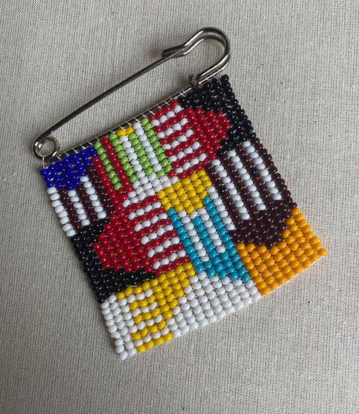 A beaded square featuring colourful geometric patterns, attached to a silver safety pin. Inspired by Contra.Joburg, the vibrant patchwork-like design bursts with colour against a neutral fabric background.