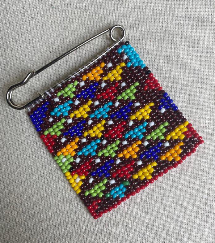 A large safety pin holds a beaded square featuring a colourful geometric pattern inspired by Contra.Joburg. The beads form zigzag stripes in red, yellow, blue, green, orange, white, and black on a light textured fabric background.
