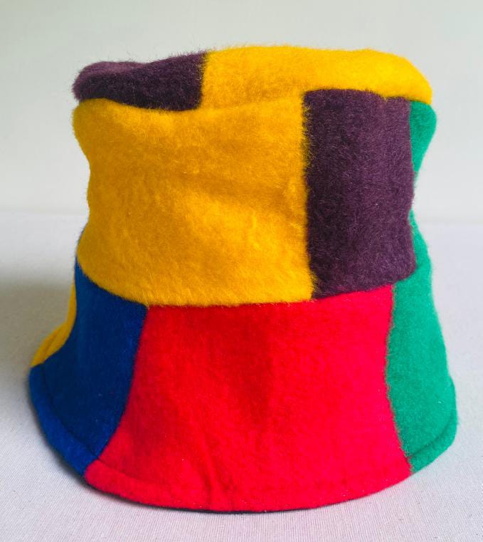 A colourful bucket hat made of soft, fuzzy fabric—reminiscent of the vibrant energy of Contra.Joburg—features large geometric patches in yellow, purple, green, blue, and red. The hat sits upright on a plain white surface with a neutral background.