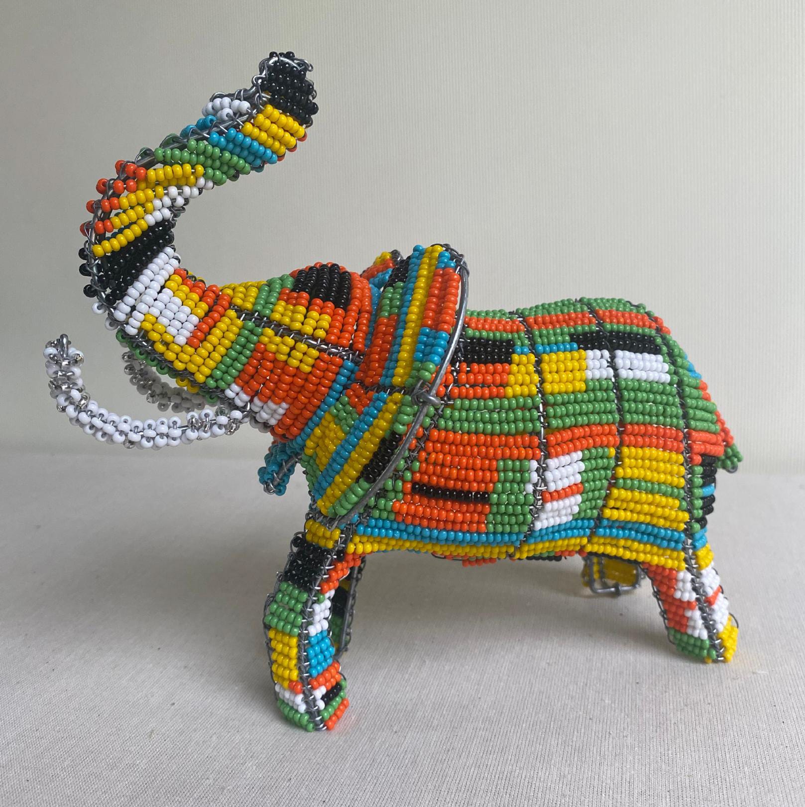A colourful beaded elephant sculpture from Contra.Joburg is shown against a plain light background. The beads form geometric patterns in vibrant shades of green, yellow, orange, red, black, and white. The elephant’s trunk is raised in a gesture of the Weight of Silence.