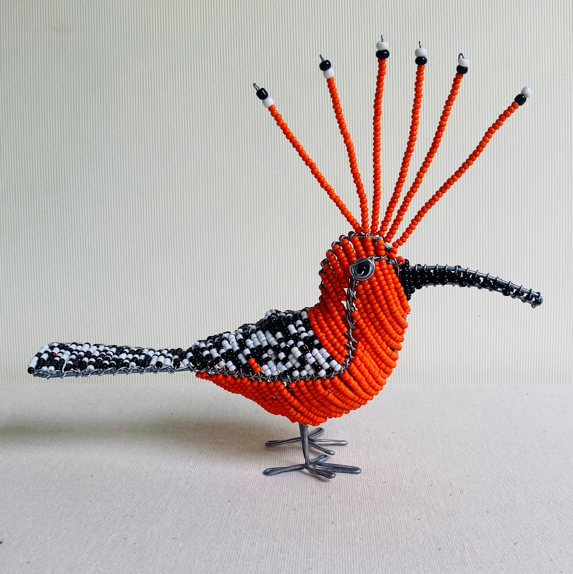 A beaded bird sculpture inspired by The Weight of Silence features a black-and-white beak, orange body, and striking wire legs. Displayed against a light striped background, this unique piece evokes the creative spirit of Contra.Joburg.