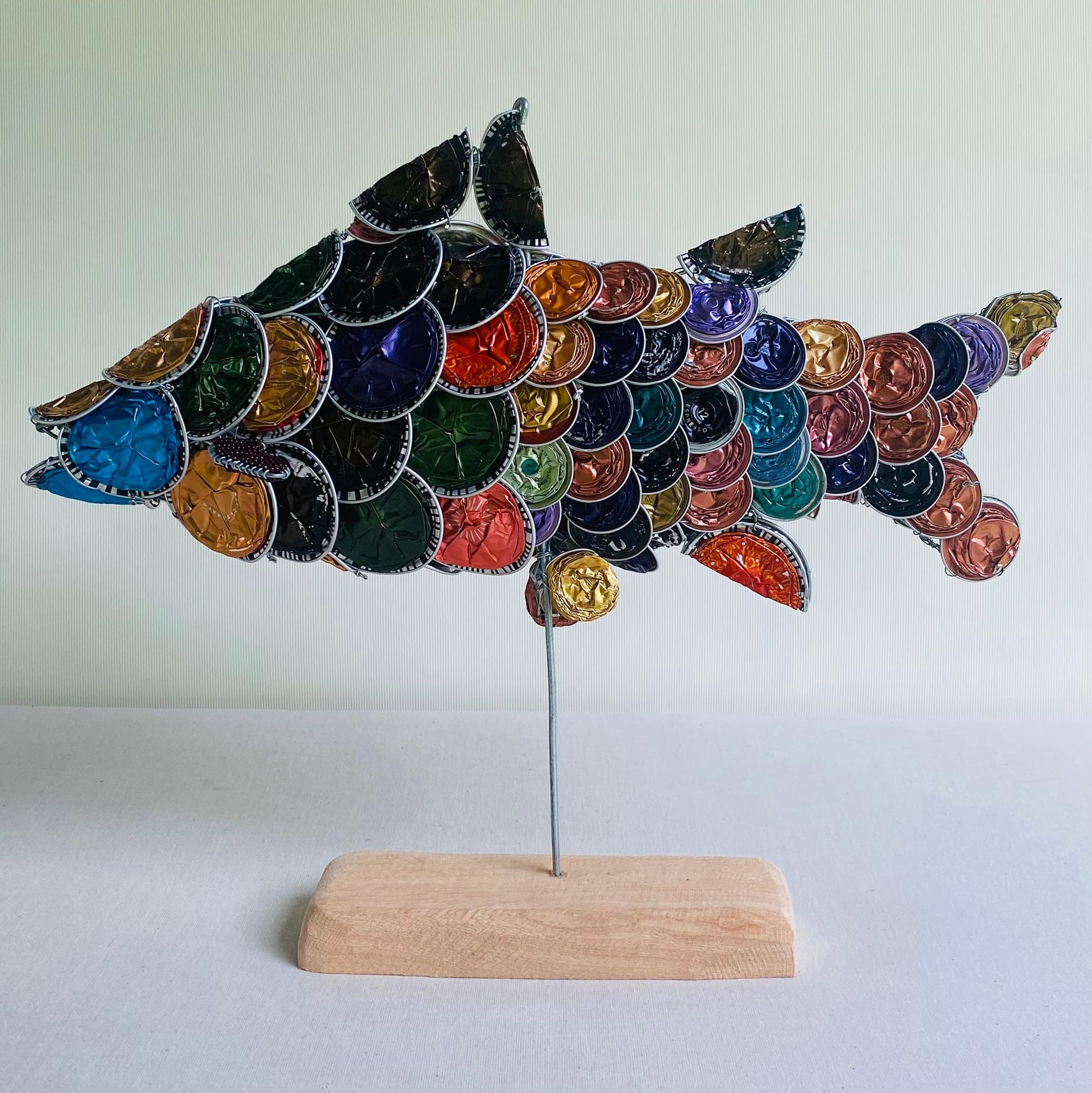 A fish sculpture made from colourful, used coffee capsules forms overlapping scales. Inspired by Weight of Silence, the capsules in rainbow hues are arranged on a thin metal rod atop a light wooden base against a pale background.