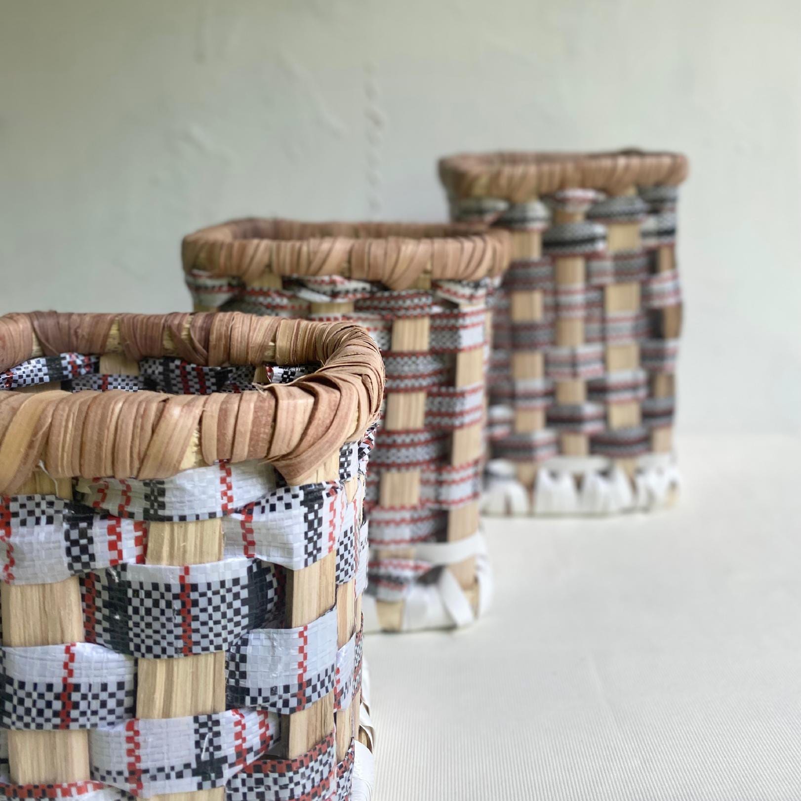 Three woven baskets with chequered patterns in natural, white, red, and black tones are arranged in a diagonal line on a light surface. Their textured, handwoven details echo the artistry of Contra.Joburg. The background is softly blurred.