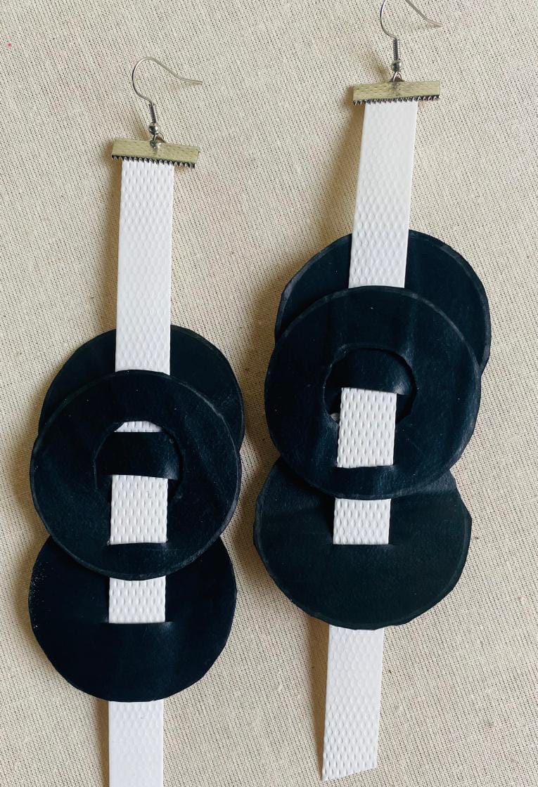 A pair of handmade earrings from Contra.Joburg features white textured vertical strips with three overlapping black velvet circles on each, capturing the Weight of Silence. The earrings have silver hooks and are displayed on a beige fabric background.