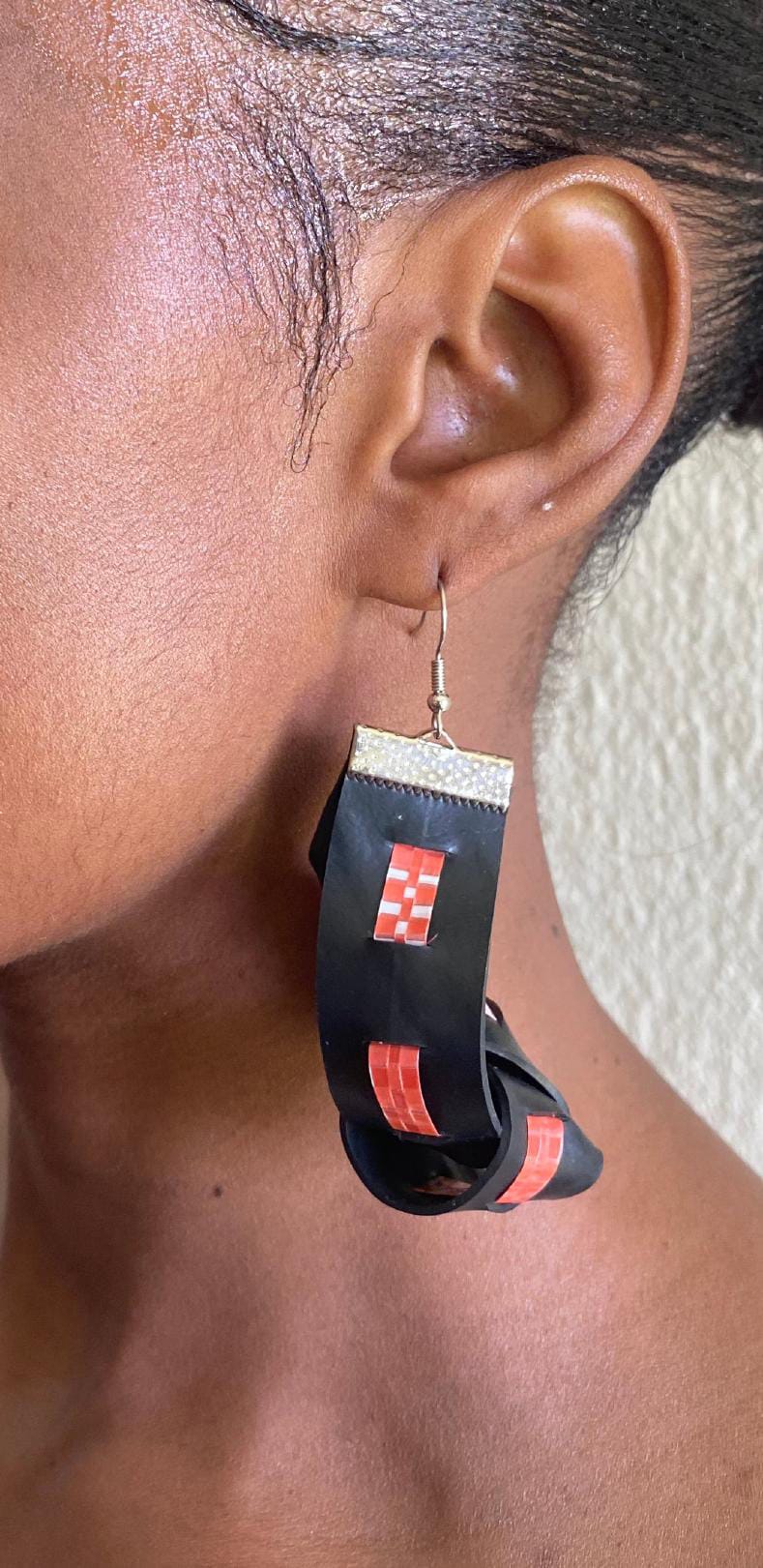 A close-up of a person's ear wearing a large, handmade black earring with red square designs and a metallic hook. Crafted from upcycled material, this Contra.Joburg piece loops in an artistic shape. The person has short hair.