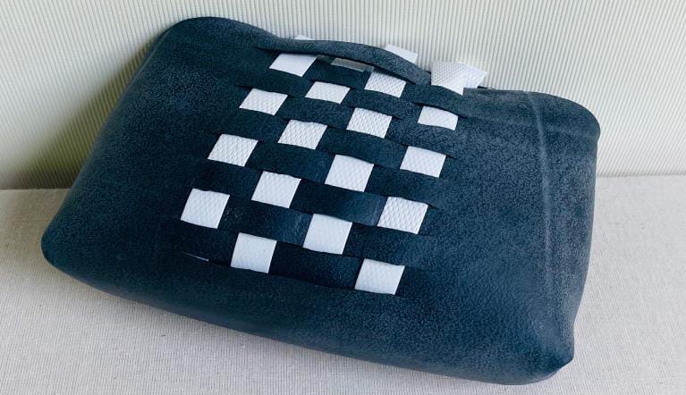 A rectangular black cushion with a grid pattern of interwoven black and white fabric strips in the centre, resting on a light-coloured fabric surface against a vertically lined light background, evokes the quiet tension of The Weight of Silence by Contra.Joburg.