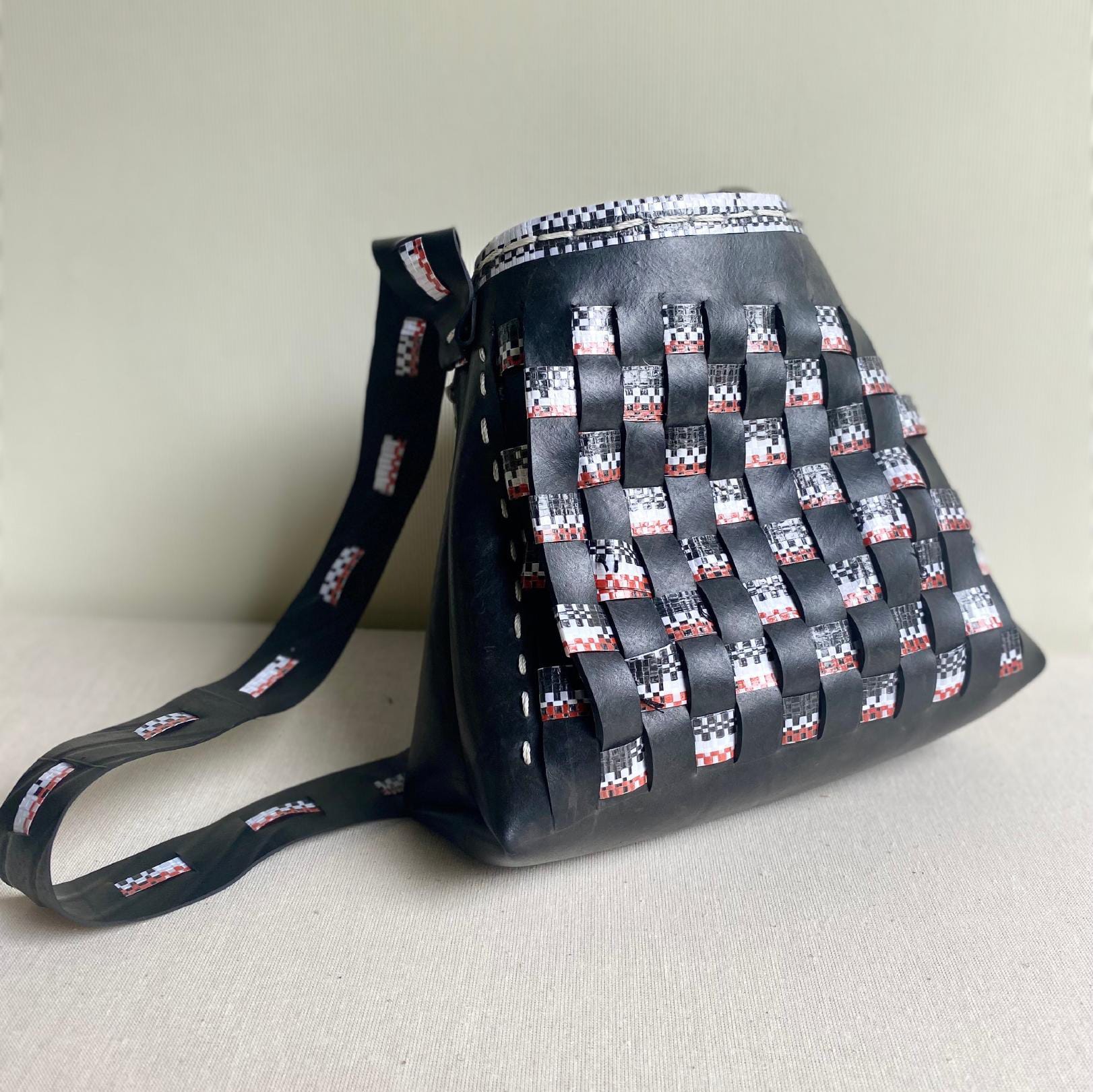 A stylish shoulder bag made of black leather with a basket-weave pattern, featuring interlaced strips of black, white, and red material. The adjustable Contra.Joburg strap displays a matching patterned design. The bag is placed against a light beige background.