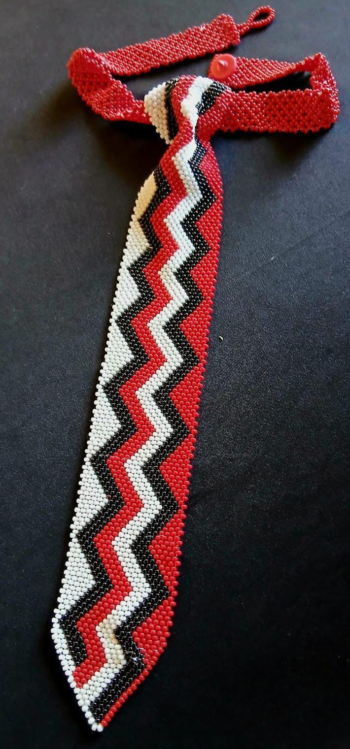 A beaded necktie with a zigzag pattern in red, white, and black lies on a dark surface. The tie tapers to a point and features a red beaded neck strap with loop and button fastening, echoing the bold style of Contra.Joburg.