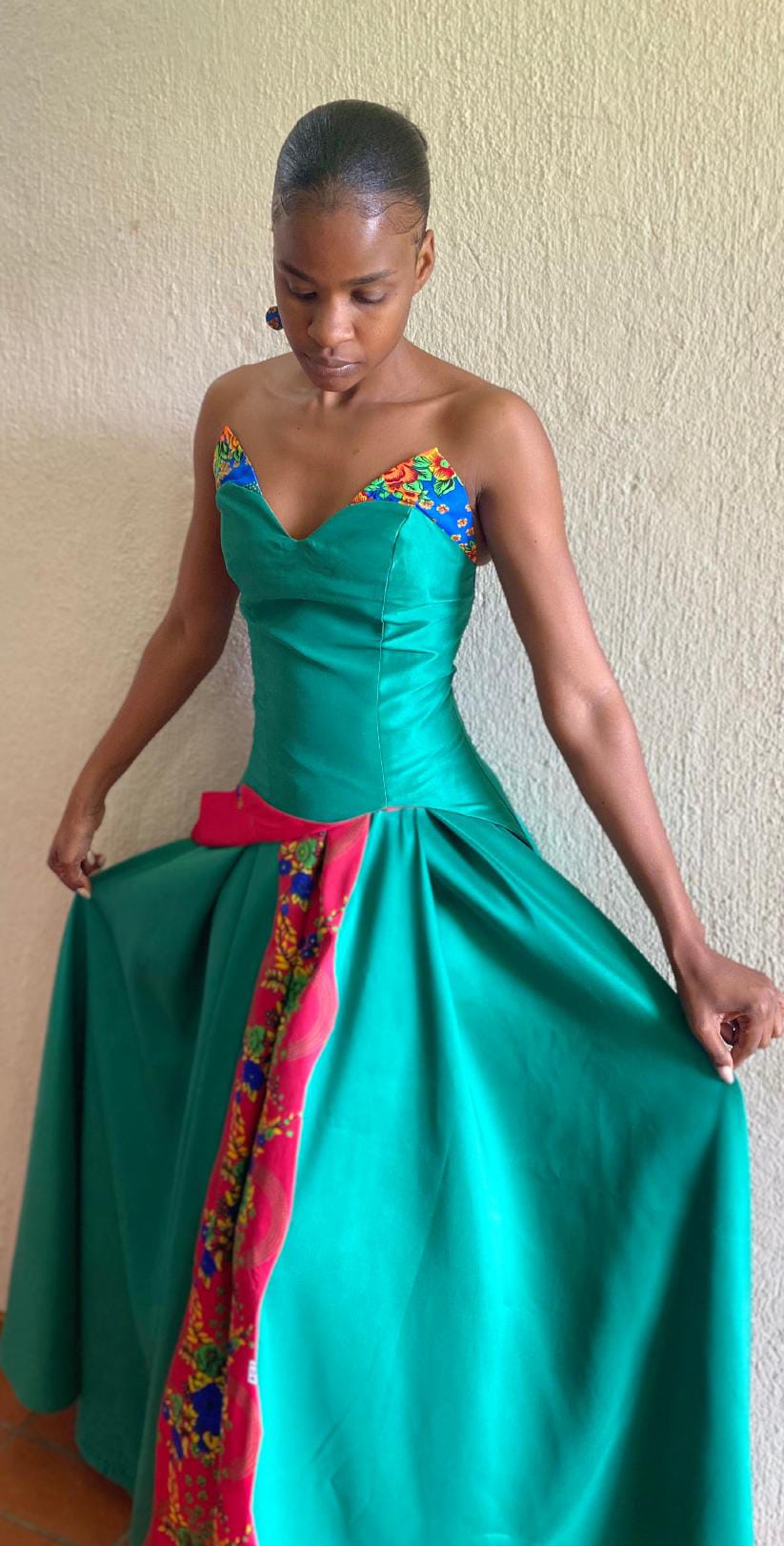 A woman in a vibrant sleeveless green dress stands against a plain, textured light wall—her attire from Contra.Joburg features colourful floral patterns on the neckline and a vertical strip, as she gently holds her skirt, embodying the Weight of Silence.