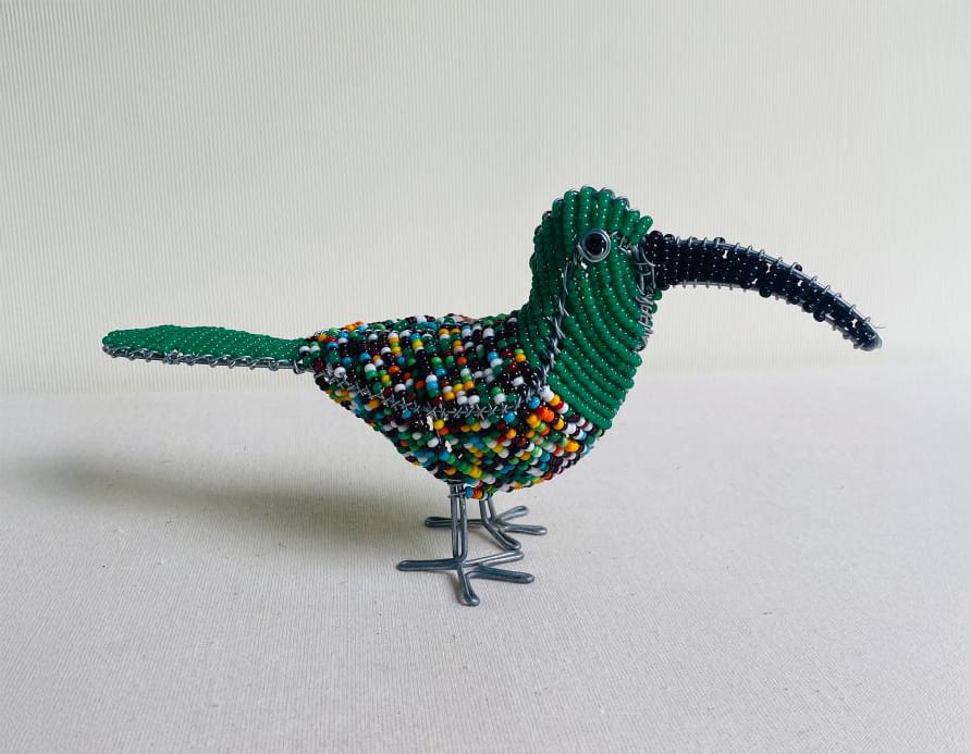 A beaded bird sculpture from Contra.Joburg, with a long, curved black beak, green head and tail, and a multicoloured body stands on wire legs against a plain background—evoking the Weight of Silence in its intricate beadwork.