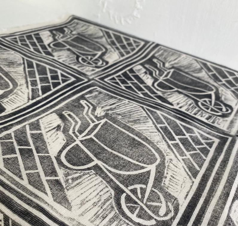Black-and-white fabric from Contra.Joburg features repeated block-print wheelbarrow designs viewed from above, with geometric patterns in the background. Draped near a white textured wall, the image highlights close-up details of this Weight of Silence print.
