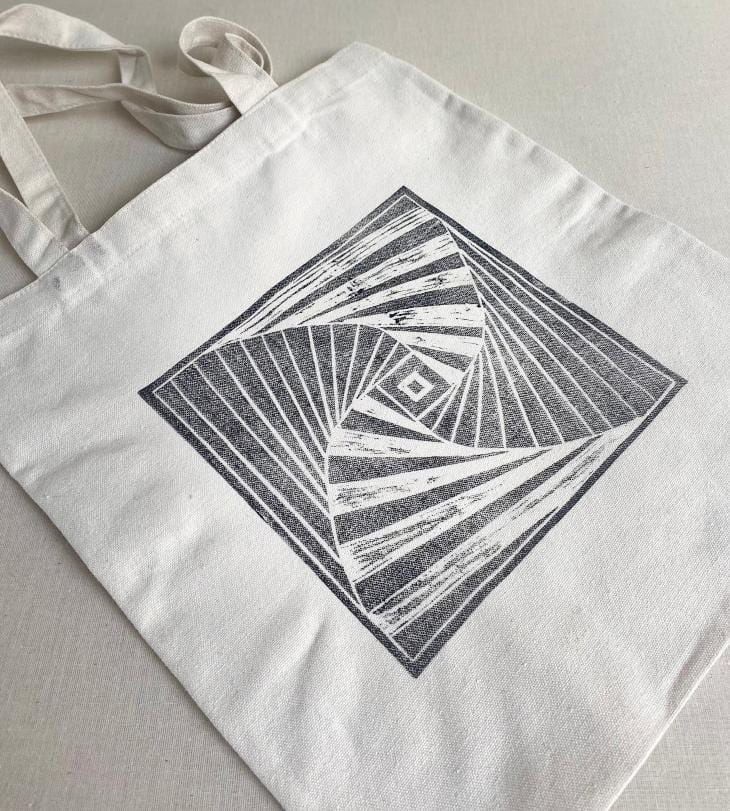 A white canvas tote bag from Contra.Joburg features a large, black geometric design—a square with swirling, radiating lines and stripes forming a hypnotic, optical illusion effect. The Weight of Silence is captured as the bag lies flat on a light, neutral surface.