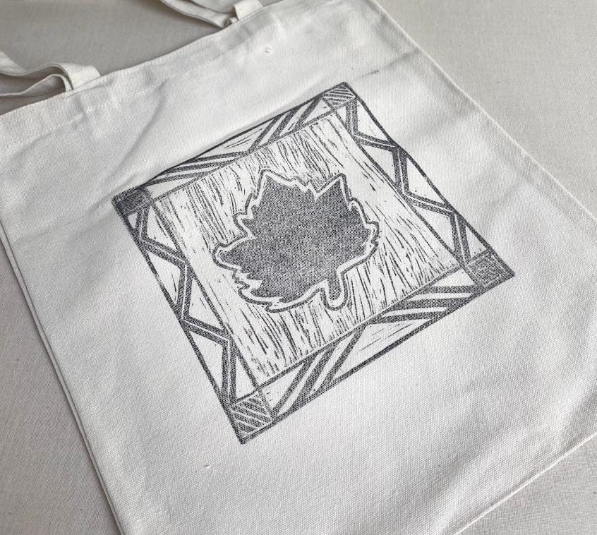 A white canvas tote bag lies flat. The bag features a black, hand-printed design of a maple leaf in the centre, framed by a square border with patterns inspired by Weight of Silence and geometric shapes from Contra.Joburg.