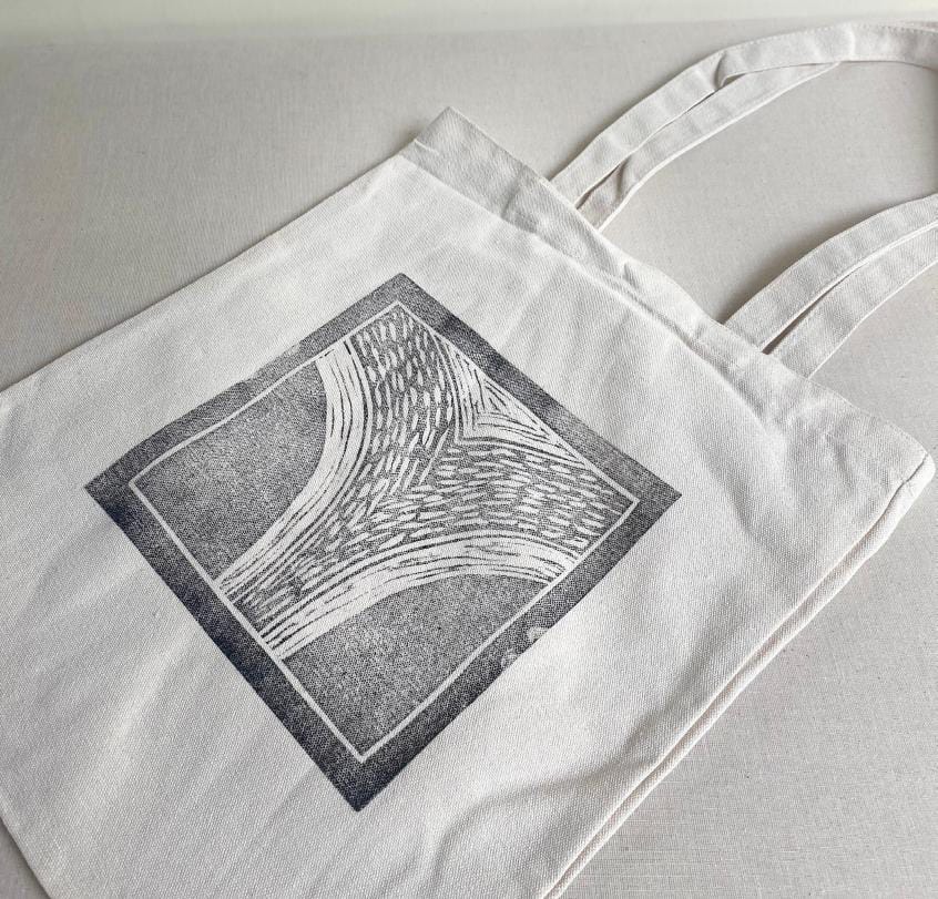 A white canvas tote bag lies on a light surface. The bag features a square black and white abstract design, inspired by Weight of Silence, resembling stylised tree branches or wood grain. The simple tote from Contra.Joburg has two handles and no other decorations.