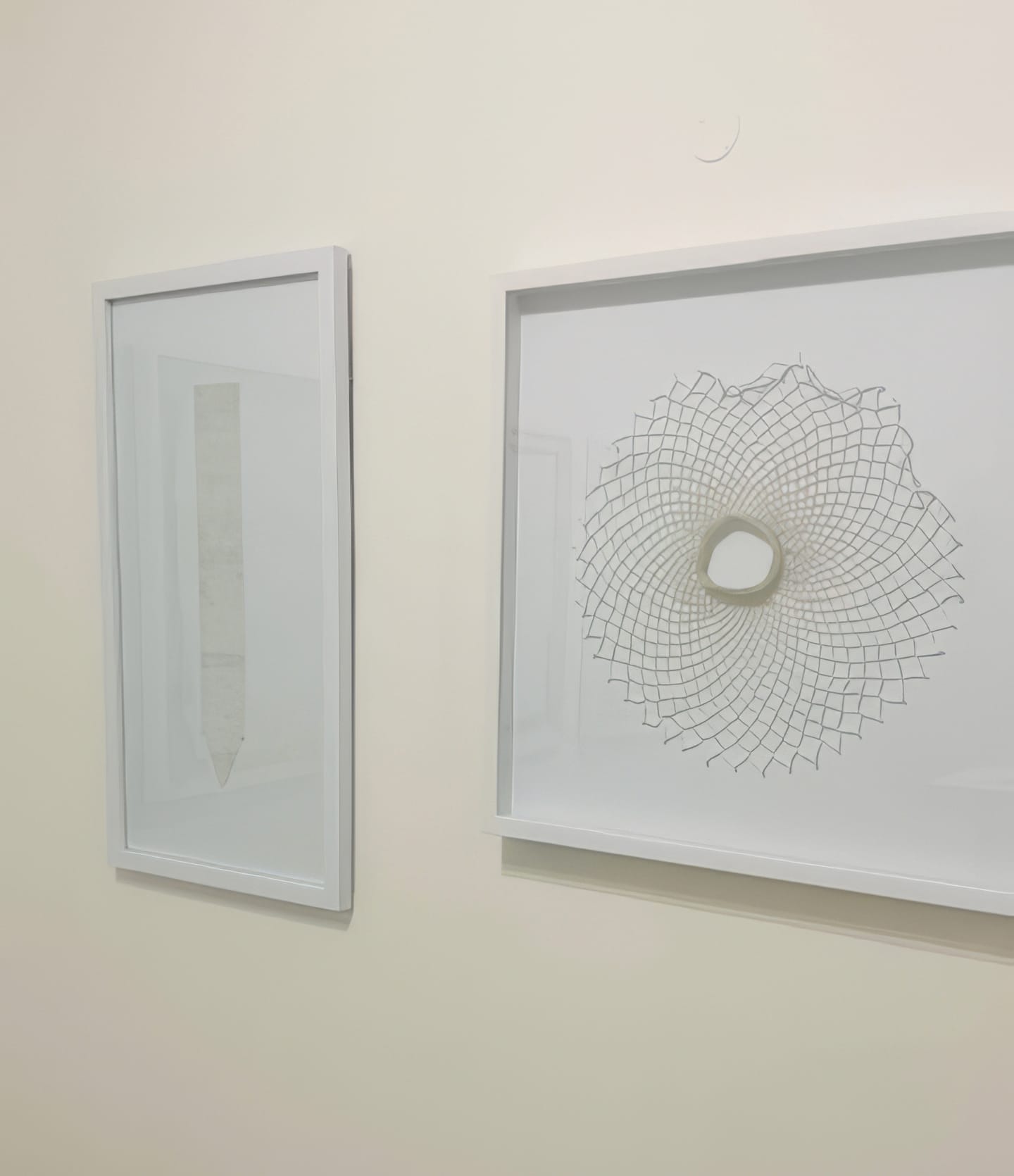 Two framed abstract artworks hang on a pale wall at Contra.Joburg. The left piece features a vertical, pencil-like shape—echoing the Weight of Silence. The right displays a spirograph pattern with an irregular central opening; both frames are white.