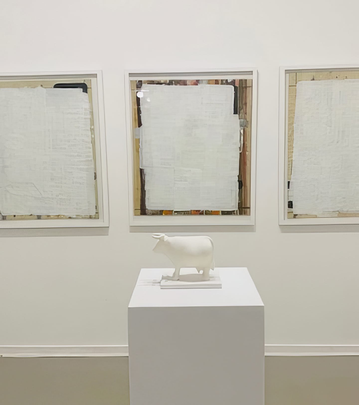 A small white sculpture of a cow, part of Contra.Joburg’s Weight of Silence series, stands on a plinth before three framed artworks—each with textured, layered surfaces that veil hidden details—all set against a plain white wall.