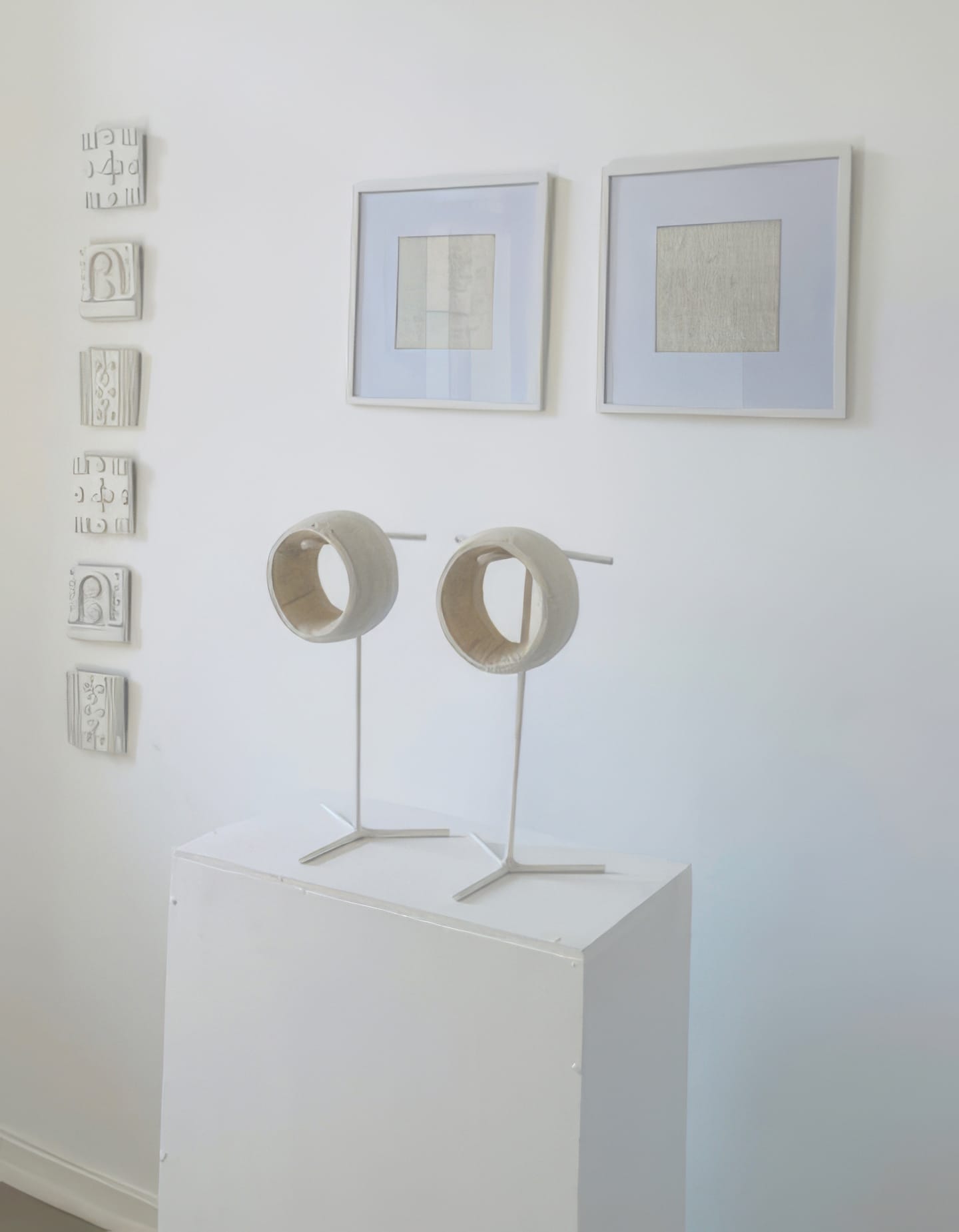 A white gallery space featuring Contra.Joburg: two circular wooden sculptures from the Weight of Silence series on metal stands atop a white plinth. Behind them, two framed abstract drawings and nine small, textured white relief panels are displayed vertically.