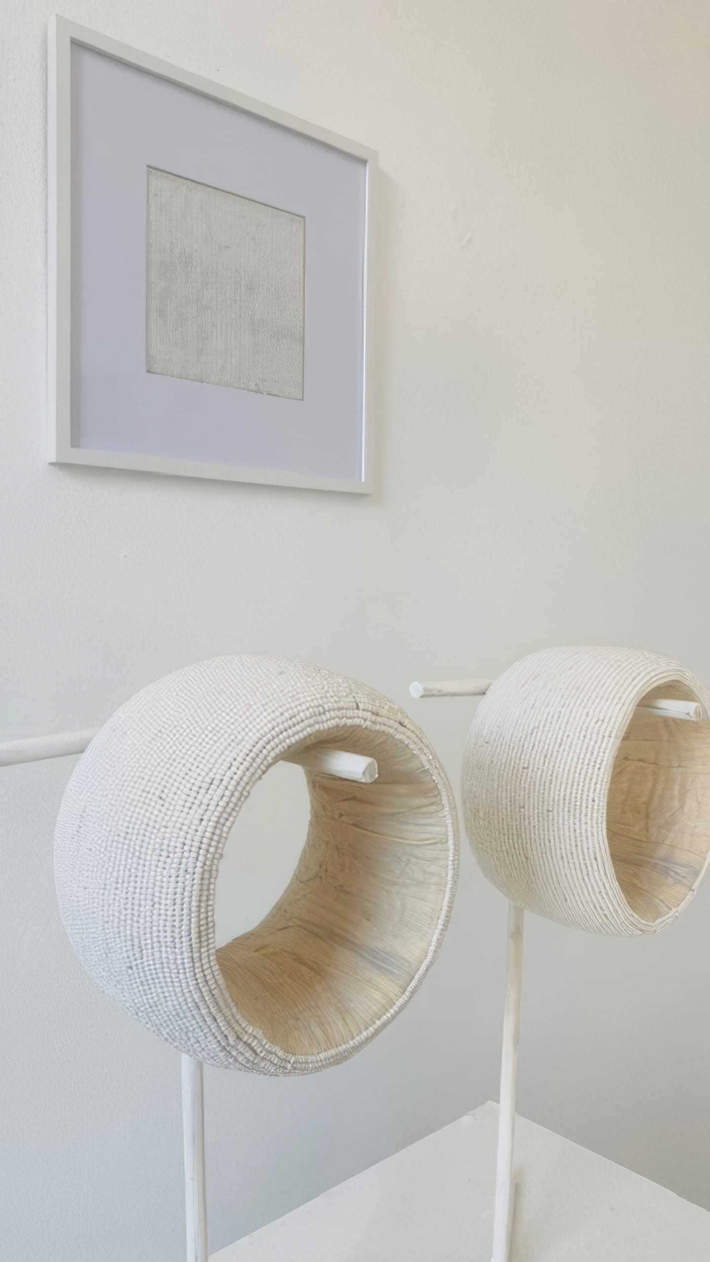 A minimal art display: two large, round, white woven sculptures from Weight of Silence rest on white stands atop a white surface. A framed abstract line drawing hangs behind them on a plain white wall, creating a calm and modern Contra.Joburg scene.