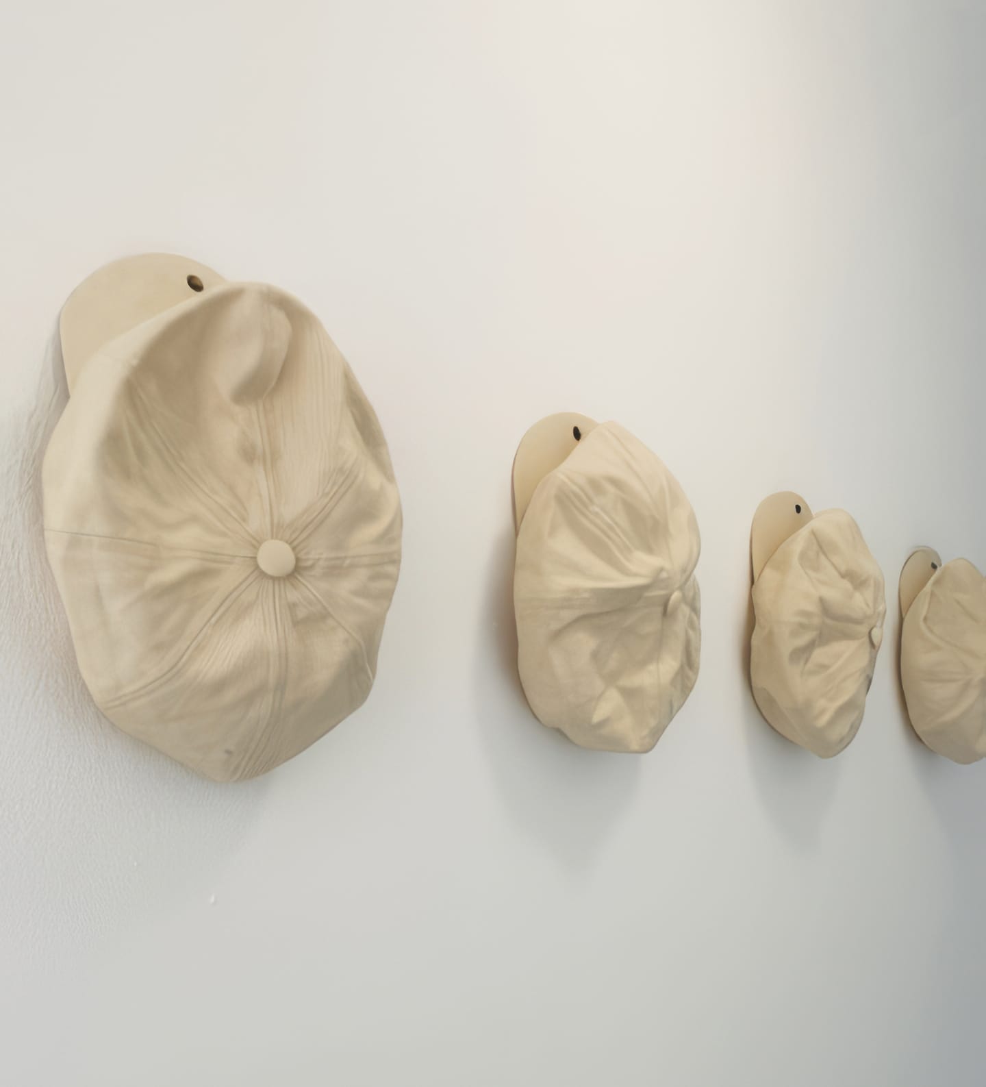 Four beige caps are neatly hung in a diagonal row on a white wall, each mounted on individual rounded hooks. Reminiscent of Weight of Silence, the identical caps create a minimalist and organised arrangement with their backs facing outward.
