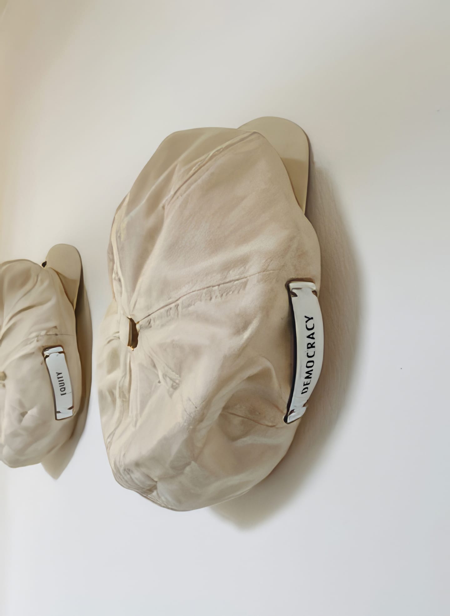Two off-white, wrinkled caps hang on wooden wall pegs against a white background. A white label with “DEMOCRACY” in black text recalls Contra.Joburg’s Weight of Silence, while the farther cap’s label remains partially visible and unreadable.