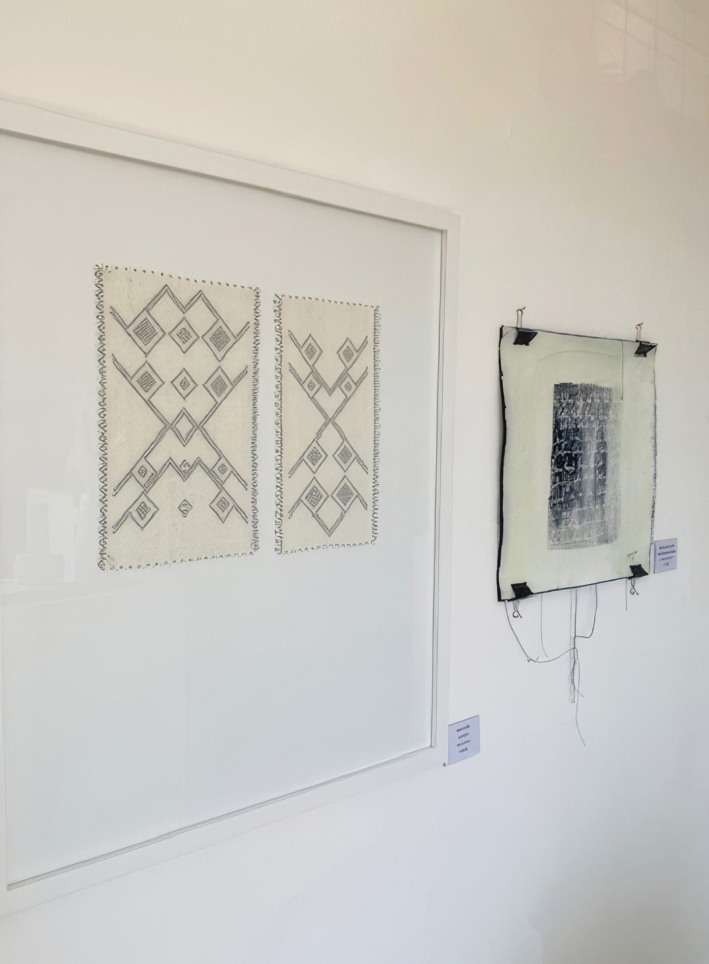 Two framed abstract artworks hang on a white wall. The left piece, Weight of Silence, features black ink diamonds and zigzags. The right artwork, part of Contra.Joburg, has a textured dark rectangle pinned to off-white fabric with loose threads.