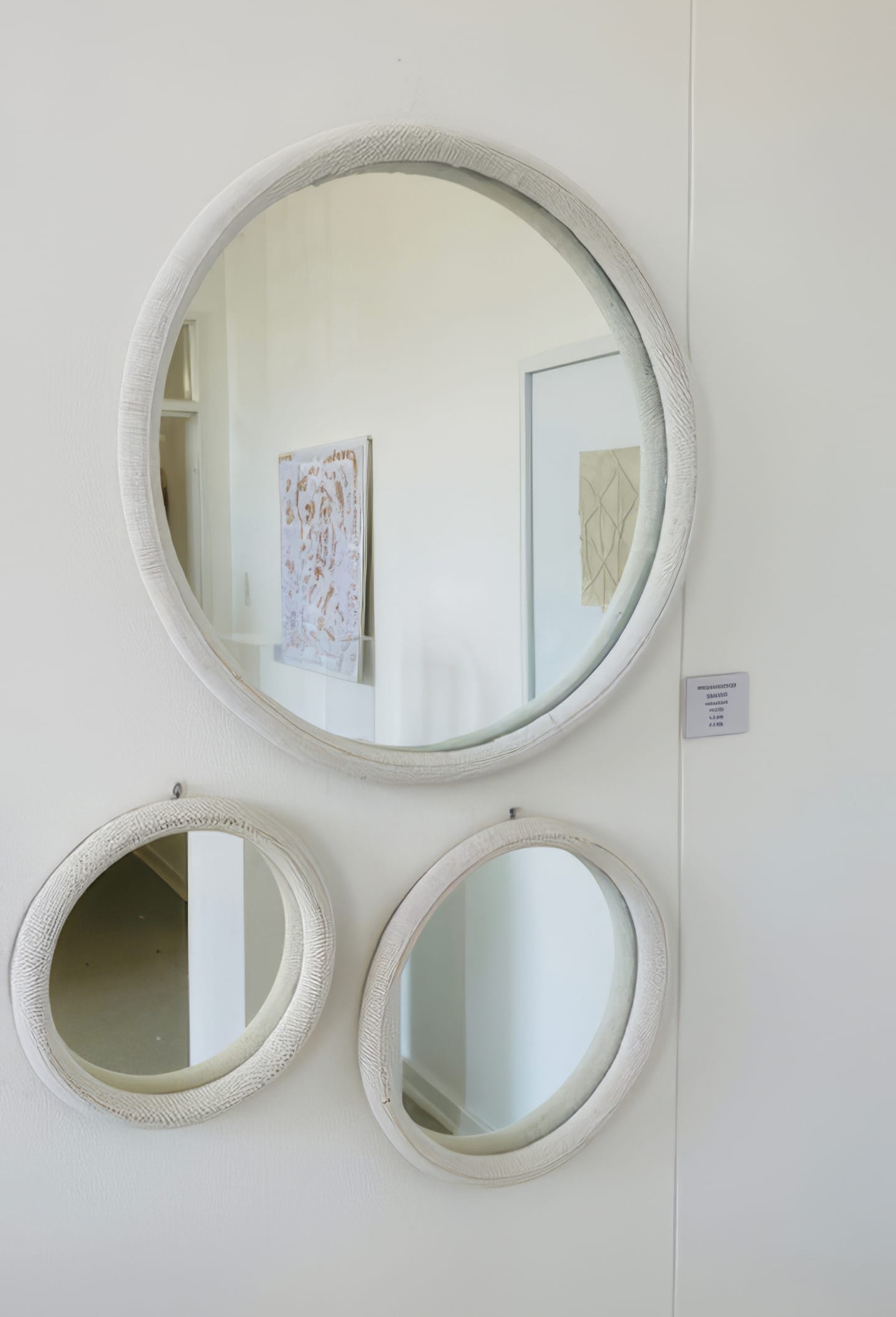 Three round mirrors with textured white frames are mounted on a white wall at Contra.Joburg. Reflections reveal a framed abstract artwork and a doorway, while a small wall label hints at an art display, possibly part of 