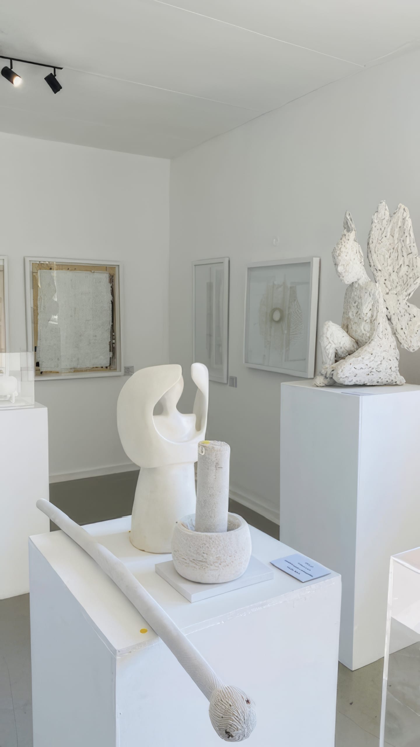 A bright, minimalist art gallery at Contra.Joburg displays white abstract sculptures on plinths—like a winged figure and a curved organic form. Framed monochrome artworks line the white walls, evoking the Weight of Silence in a serene, modern atmosphere.