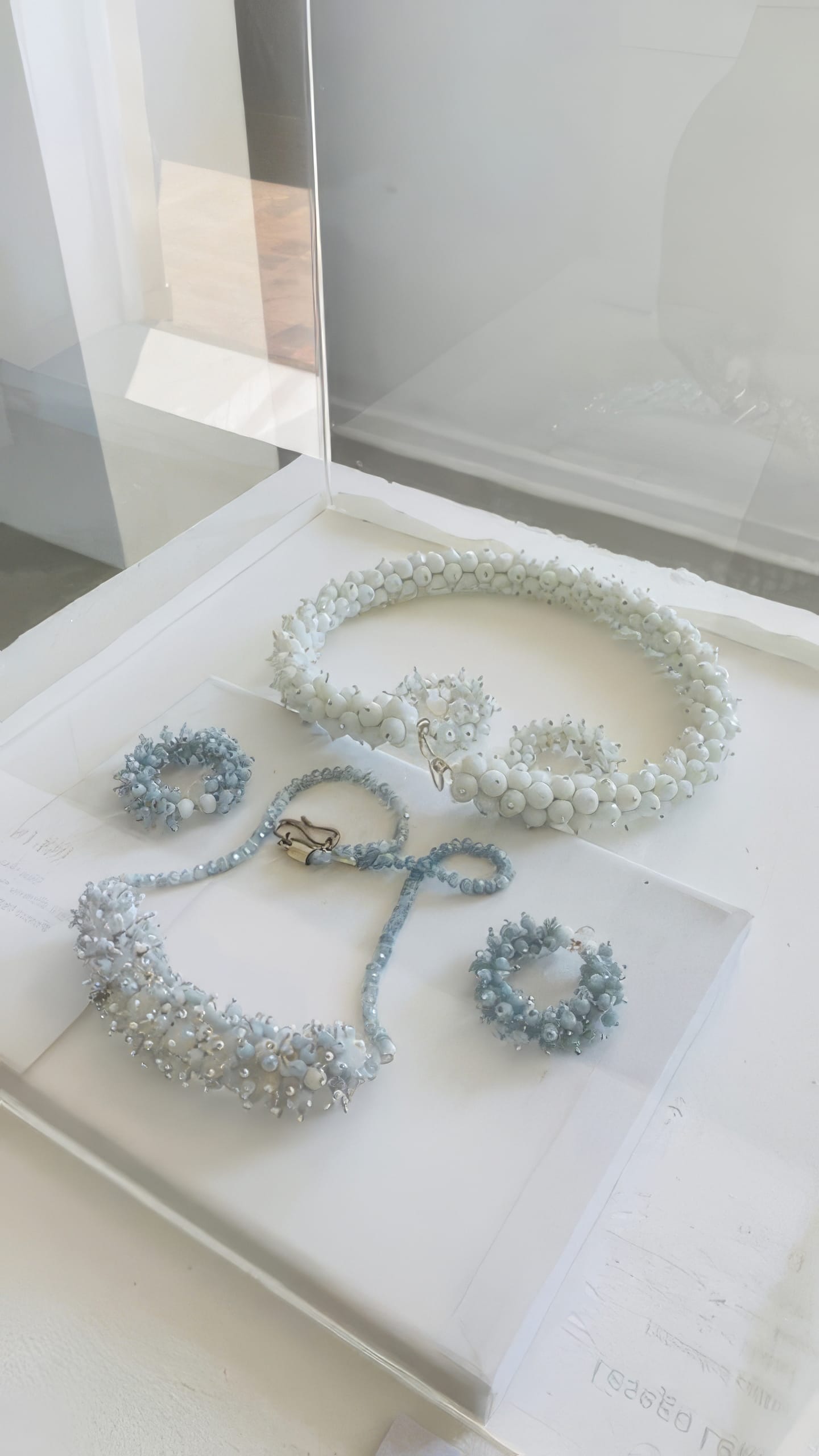 A jewellery display case with a necklace, bracelet, ring, and earrings from Contra.Joburg. Each piece features white and pale blue beads and pearls, creating a delicate floral texture. The clean display is set by a sunlit window, evoking the Weight of Silence.