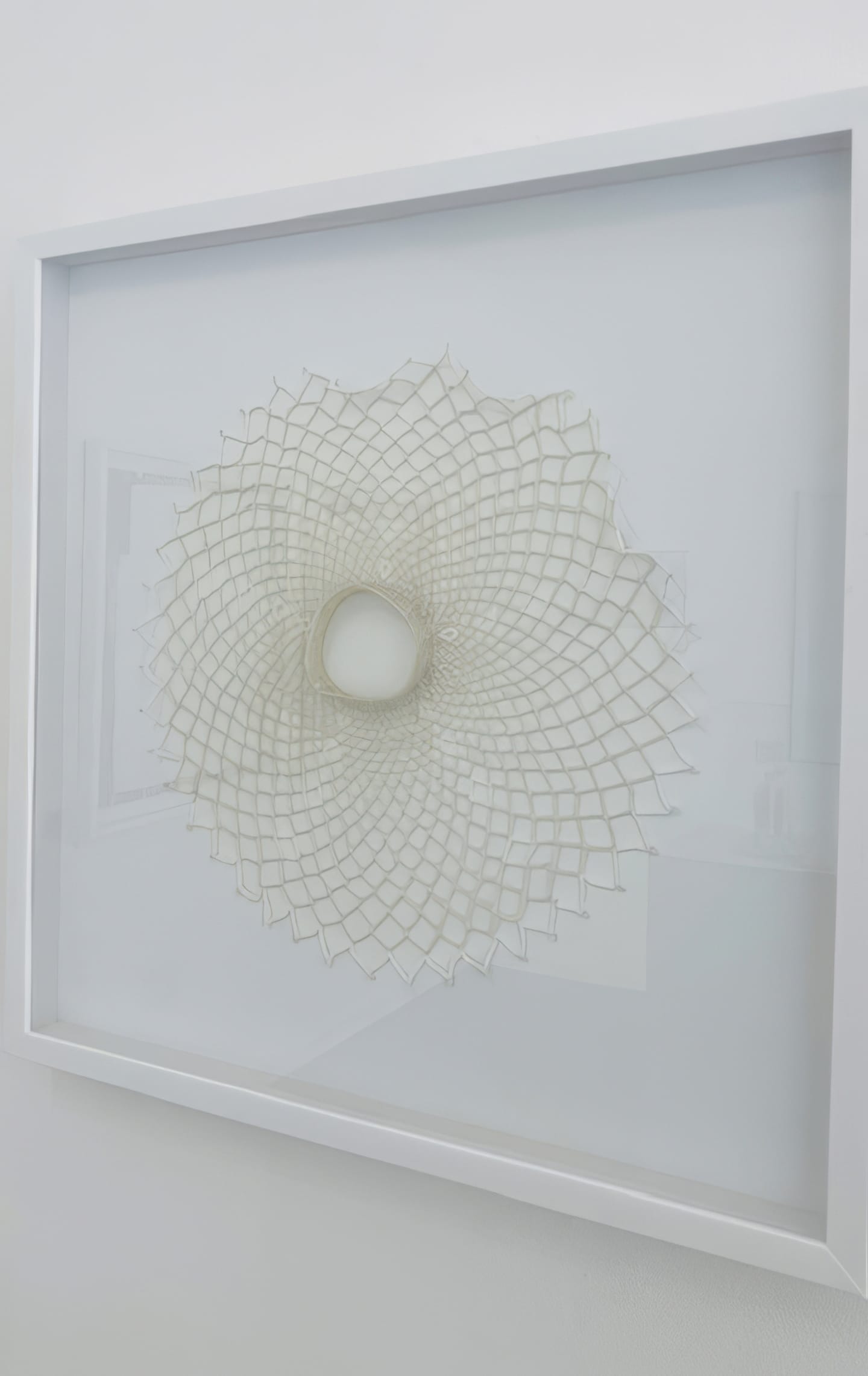 A white framed artwork hangs on a light wall at Contra.Joburg, featuring a delicate, three-dimensional geometric pattern—resembling the Weight of Silence—radiating outward with intricate, lattice-like details centred around a hollow circular opening.