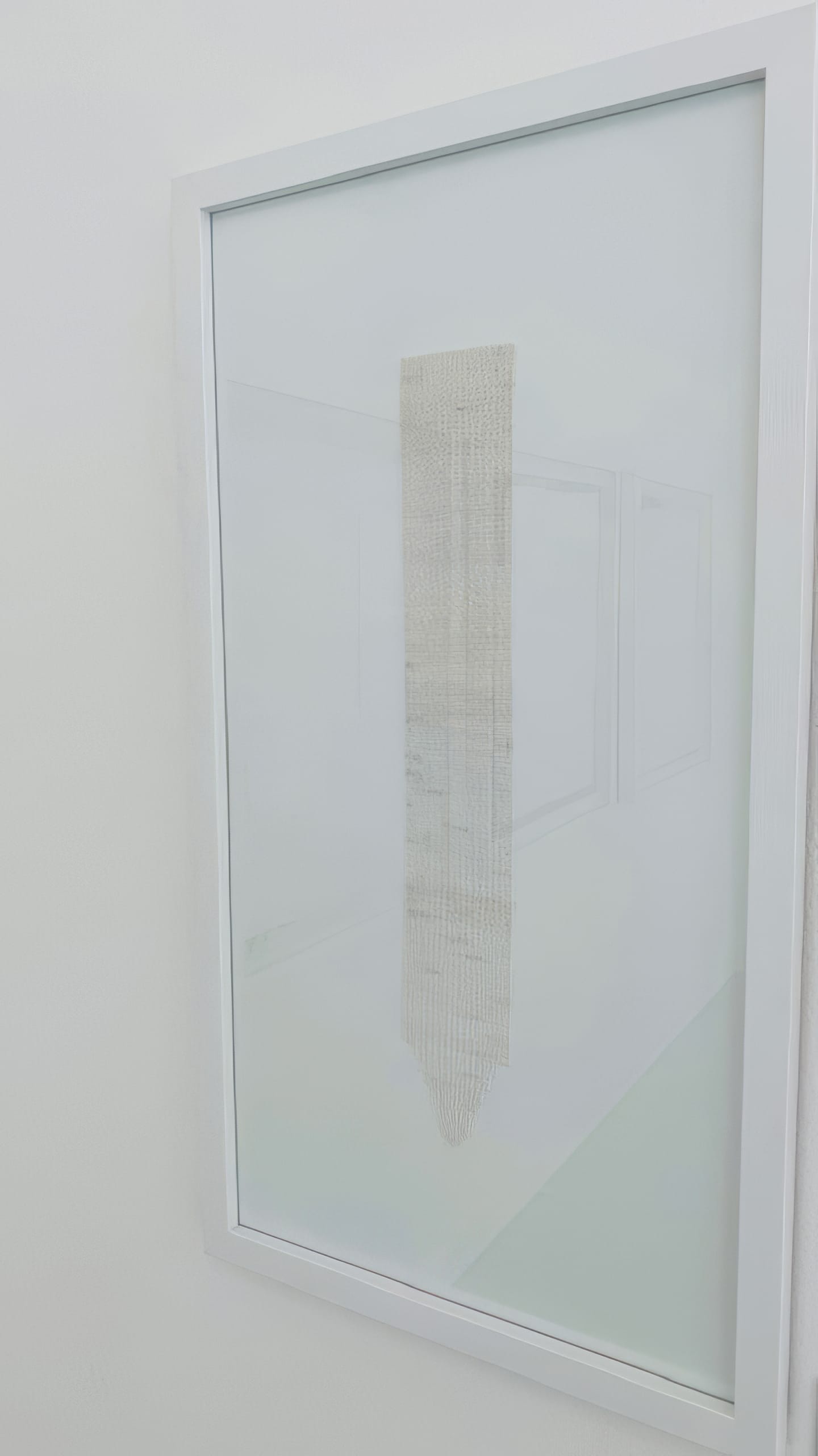 A white-framed glass case displays a narrow, vertically hung, textured textile piece in light beige tones. The minimalist background and faint reflections highlight the Weight of Silence, emphasising the simplicity and delicacy of the artwork.