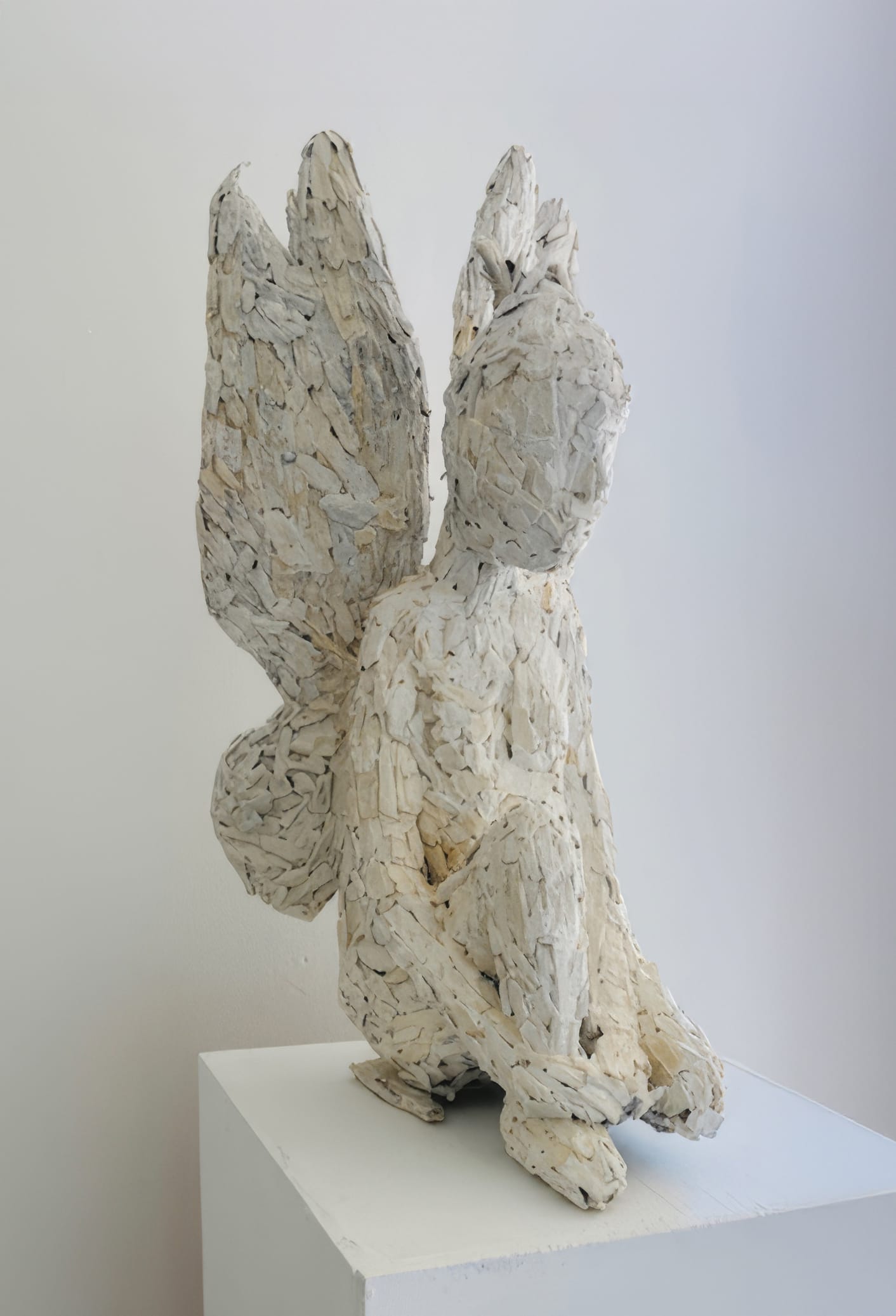 A textured sculpture of a winged, seated figure—titled *Weight of Silence*—is formed from layered, off-white fragments. This abstract piece, shown at Contra.Joburg, rests quietly on a white plinth against a plain, light background.