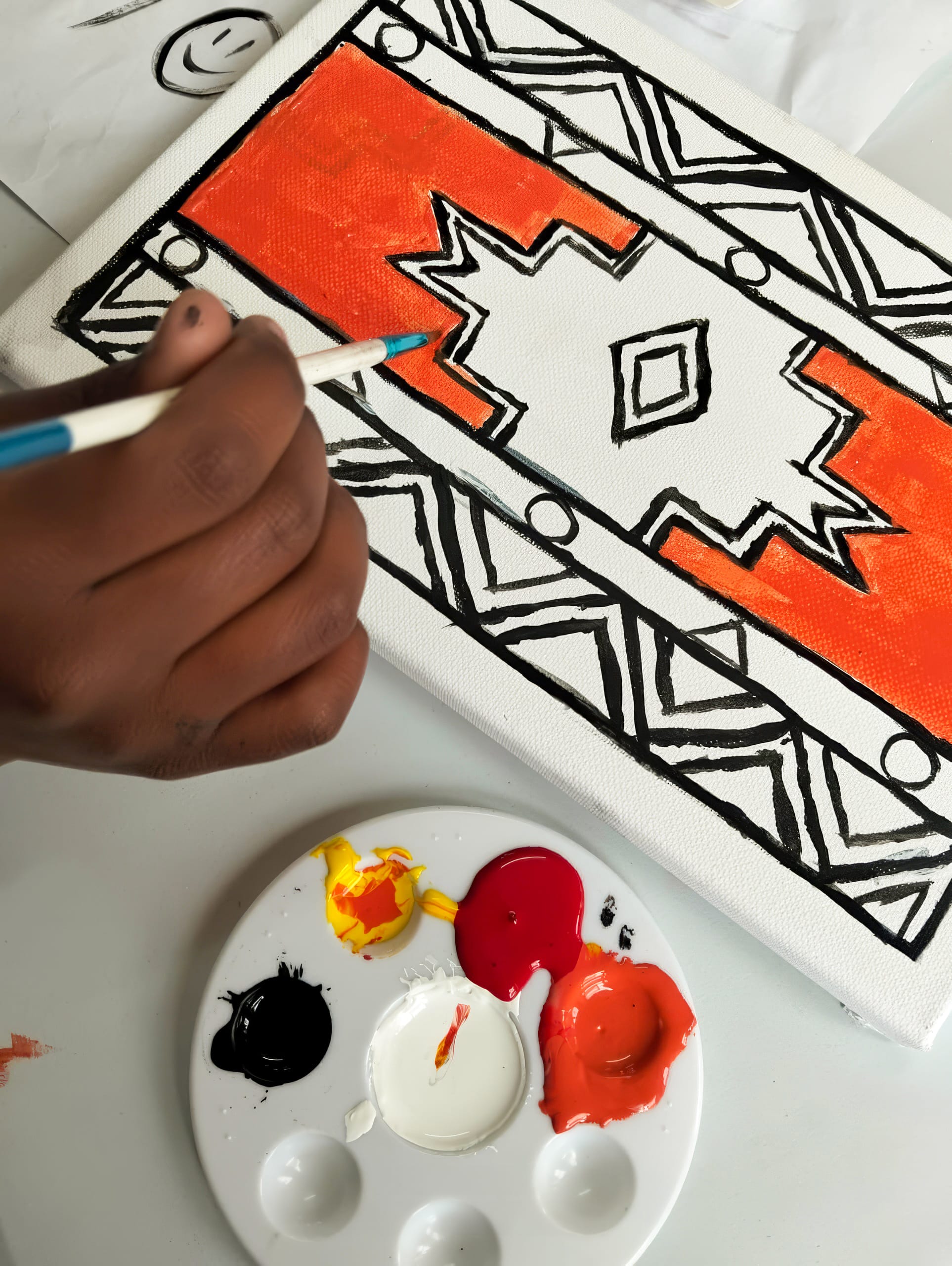 A hand uses a paintbrush to fill bright orange paint within a geometric tribal pattern on a white canvas, celebrating the bold black lines and symmetrical shapes. This vibrant design is inspired by the Legacy Project of Gogo Mahlangu.