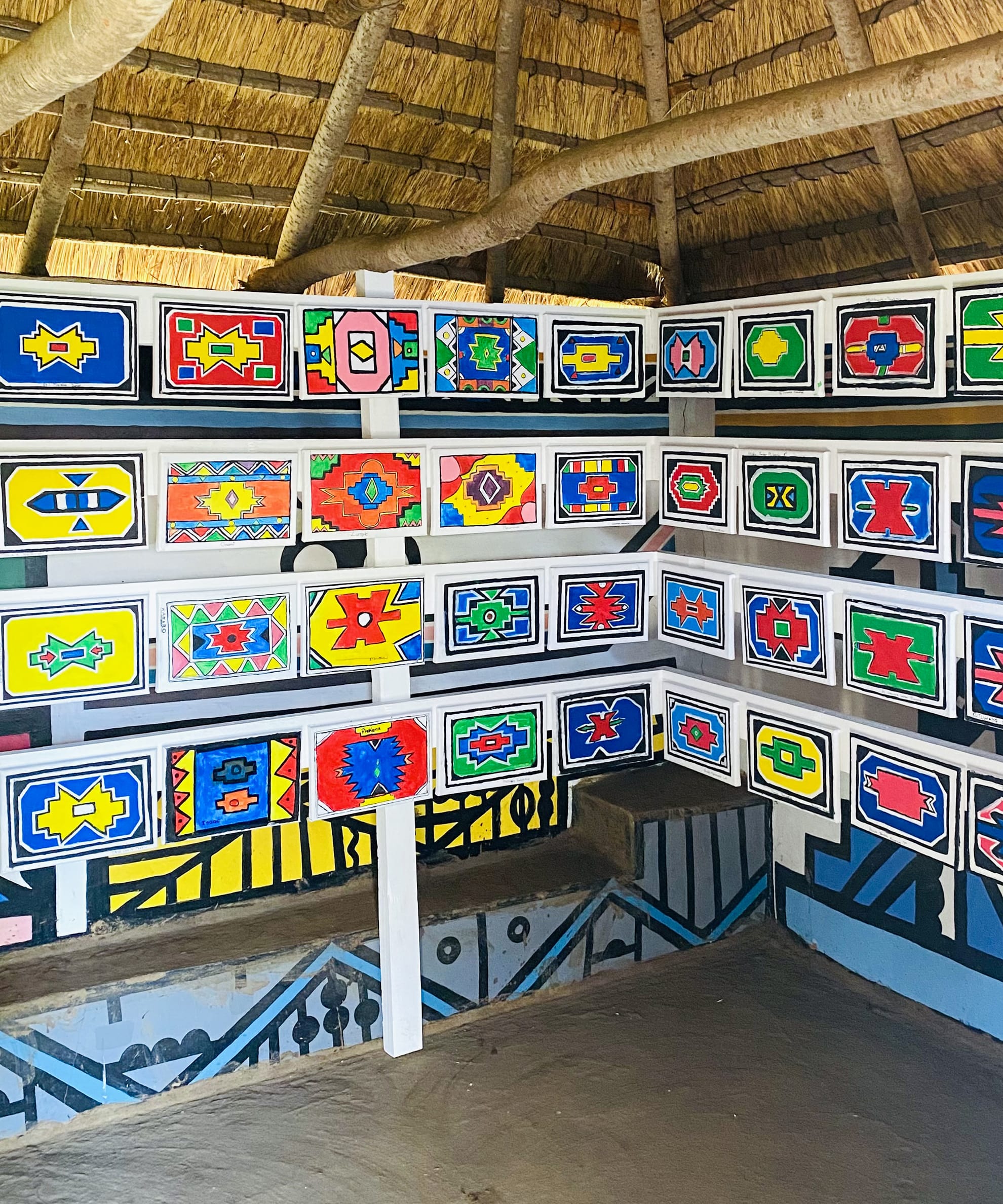 Colourful geometric Ndebele art covers interior walls of a round, thatched-roof hut. Bold patterns fill neat rows, celebrating the traditional South African design and honouring Gogo Mahlangu’s legacy project in every vibrant panel.