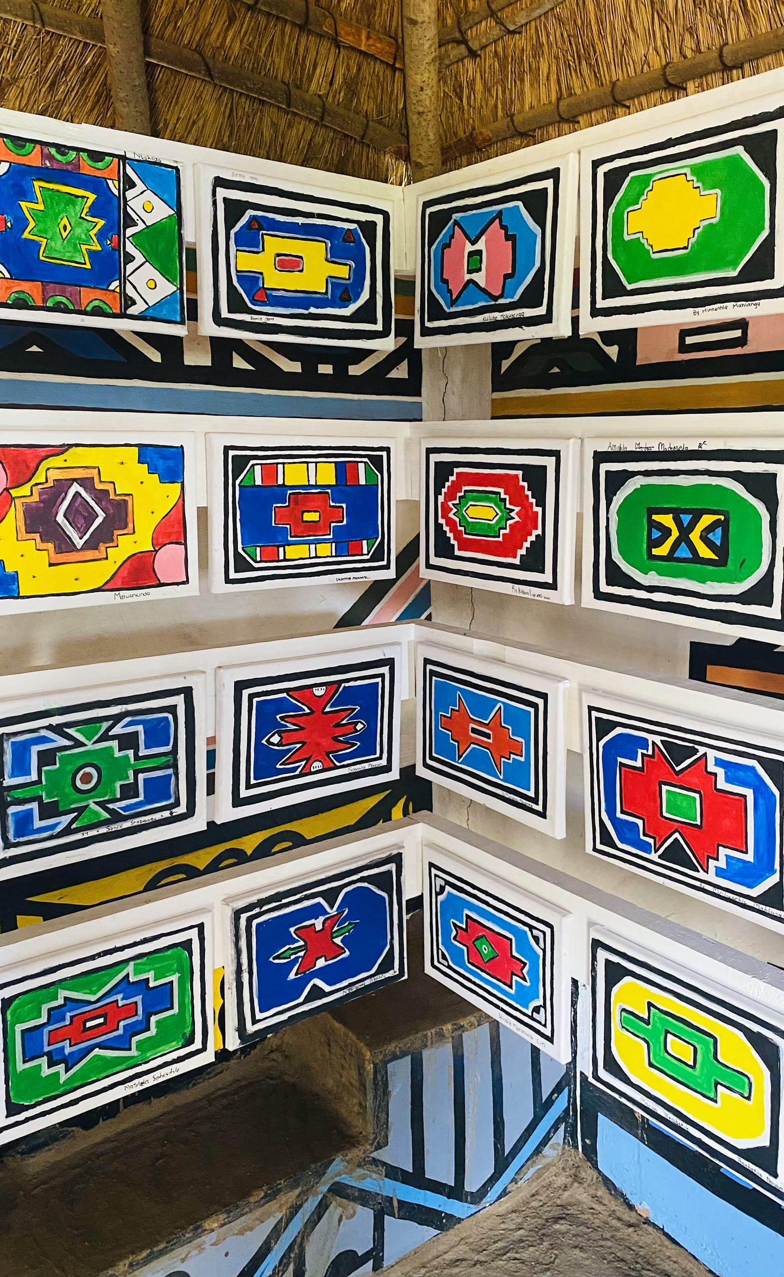 A corner of an interior hut with white walls features colourful, geometric, rectangular Ndebele patterns in red, yellow, blue, green, and black—a vibrant nod to Gogo Mahlangu and her Legacy Project. Built-in mud benches sit below visible thatched roof beams.
