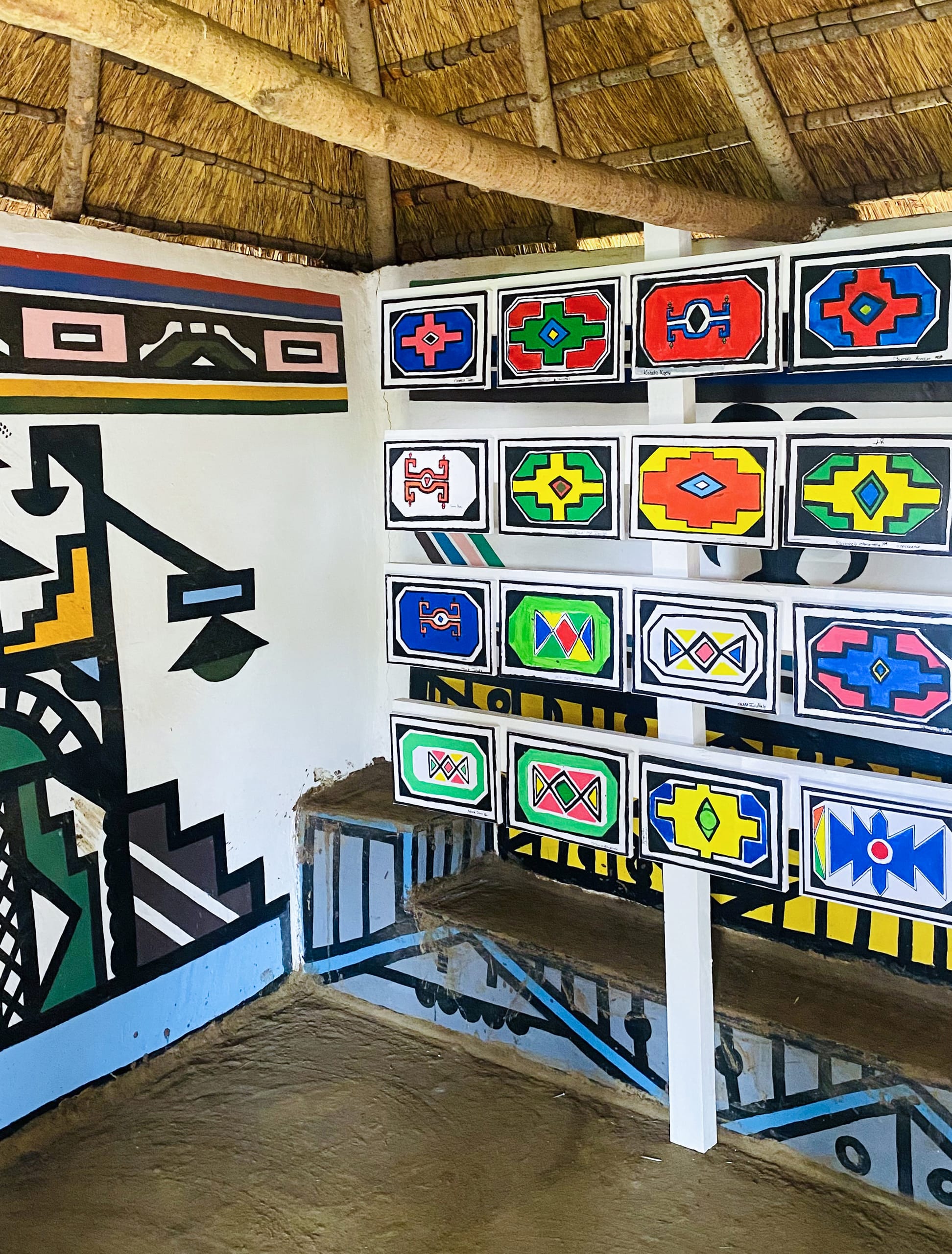 A display of colourful, geometric Ndebele wall art lines the hut, celebrating Gogo Mahlangu’s legacy project. The bold patterns in black, blue, green, yellow, and red adorn the walls beneath a thatched roof and above a mud floor.
