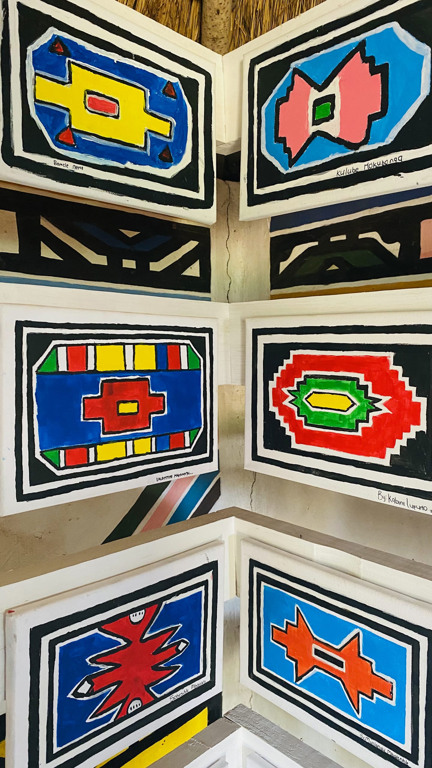 Six rectangular, brightly coloured geometric paintings are displayed on a white wall. Inspired by African traditional art, this Celebration is part of the Gogo Mahlangu Legacy Project, with labels beneath each bold, symmetrical artwork.