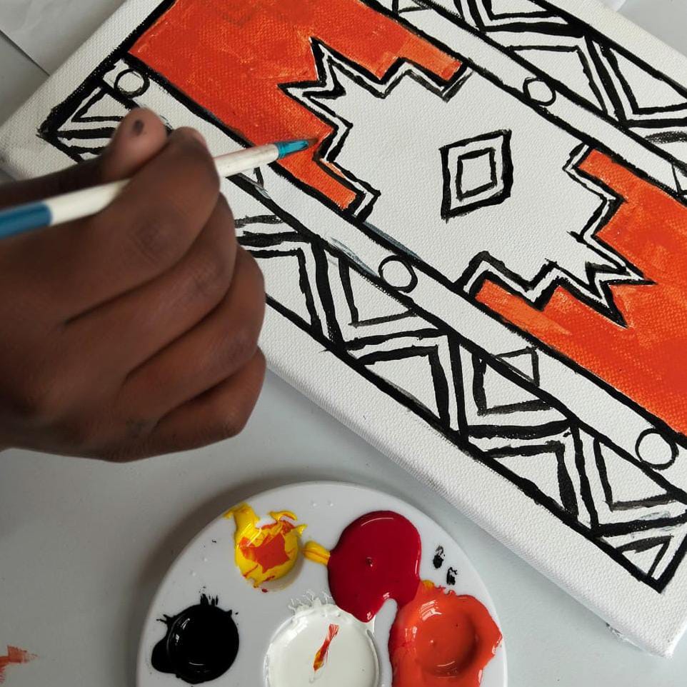 A hand holds a paintbrush, filling in an orange geometric pattern on a canvas. The design, inspired by tribal motifs, pays tribute to the Gogo Mahlangu Legacy Project and features bold black outlines and intricate shapes—a true celebration of heritage.