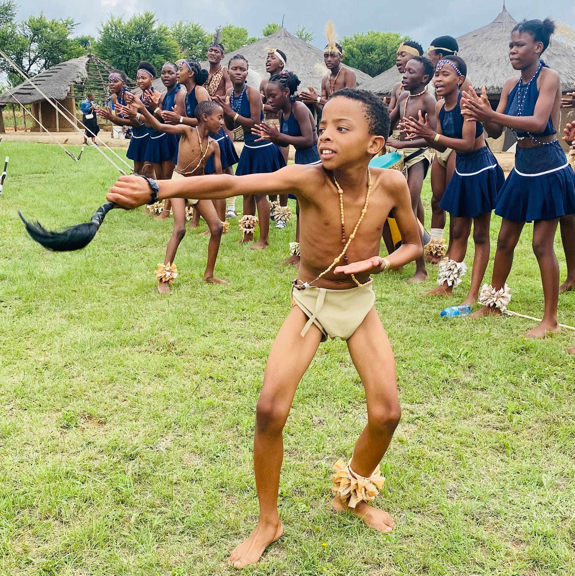 A barefoot boy in traditional attire dances energetically, holding a horn, as part of the Legacy Project celebration, whilst girls in navy dresses and beaded jewellery clap and sing behind him on a grassy field with thatched huts and trees in the background.