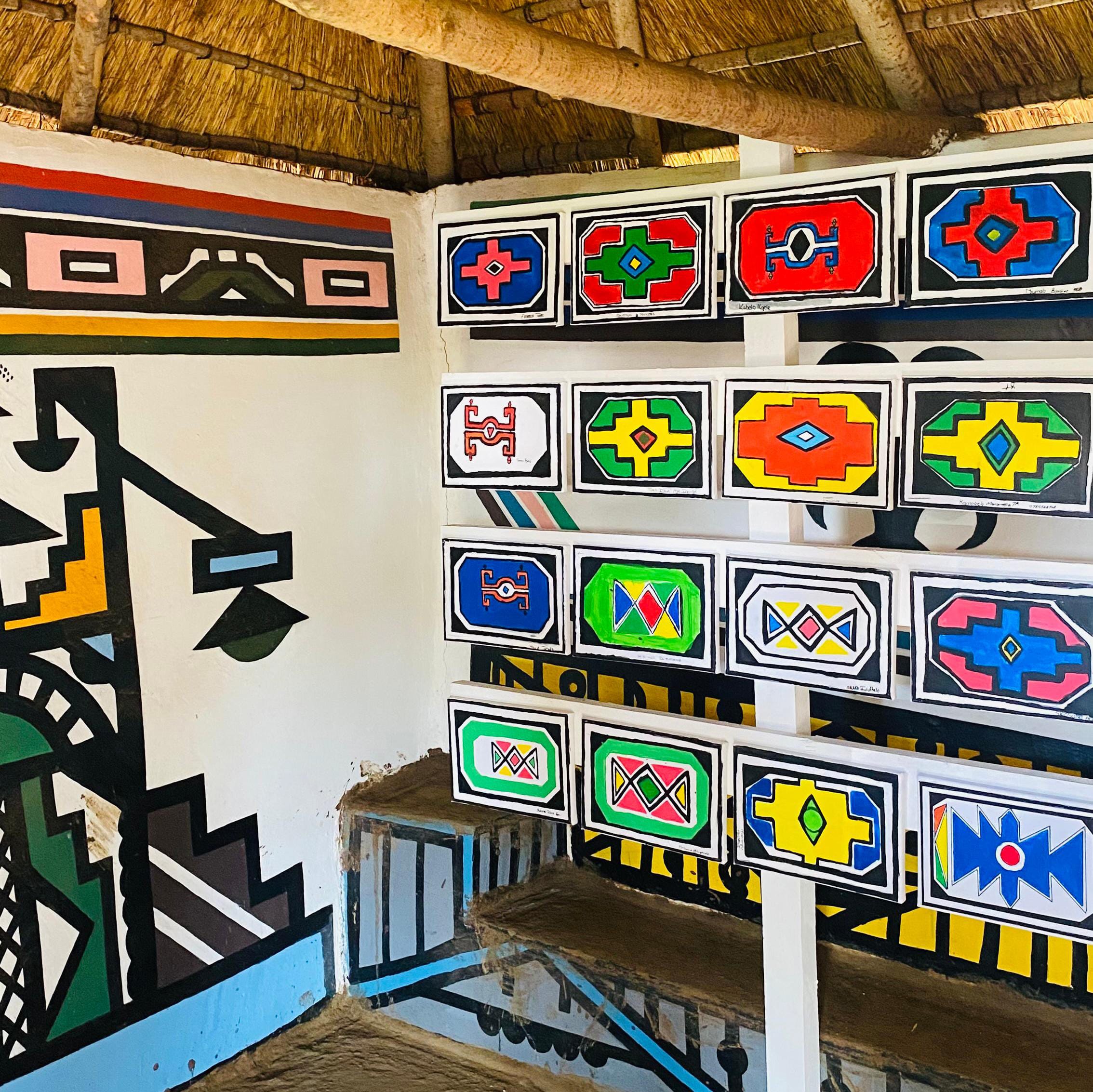 A room with a thatched roof features bold geometric mural art and colourful rectangular patterns, each with intricate designs in vibrant hues, celebrating traditional African Ndebele artwork and honouring the Legacy Project of Gogo Mahlangu.