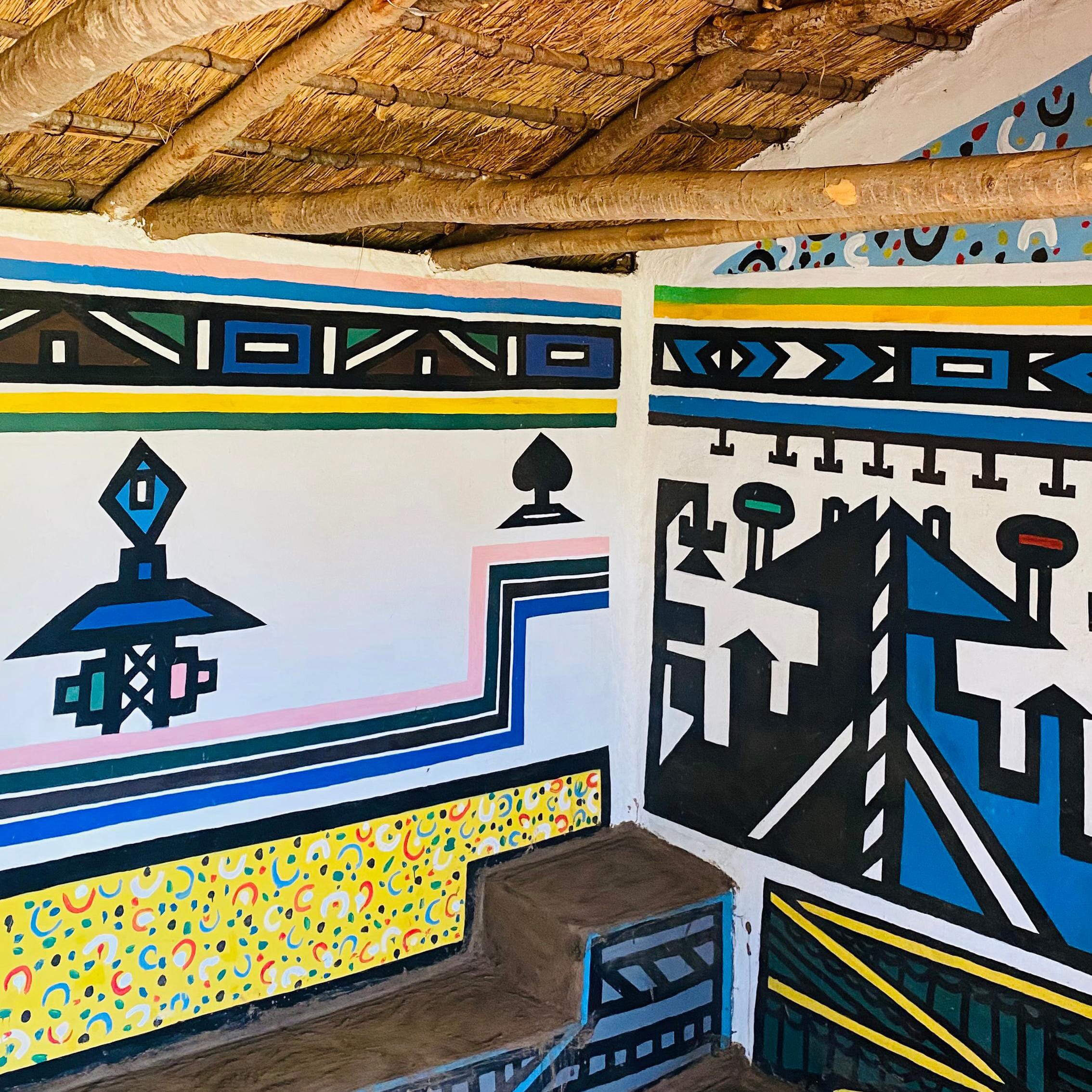 A room with colourful, geometric Ndebele mural art covering the walls celebrates Gogo Mahlangu’s Legacy Project, featuring bold lines, shapes, and patterns. The woven thatch ceiling is supported by rustic wooden beams.