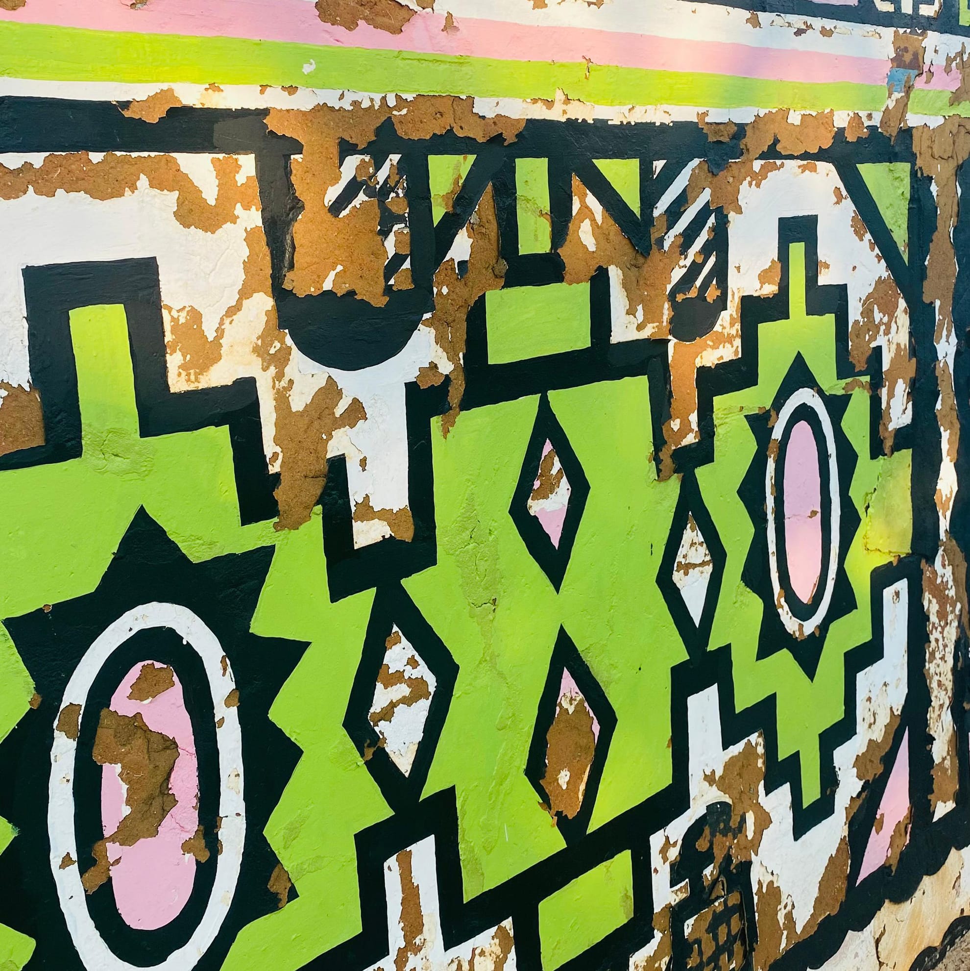 A heavily weathered wall mural, part of the Legacy Project, features bold geometric patterns in lime green, black, white, and pink. Large patches of peeling brown paint reveal the underlying surface, giving this celebration of art a textured appearance.