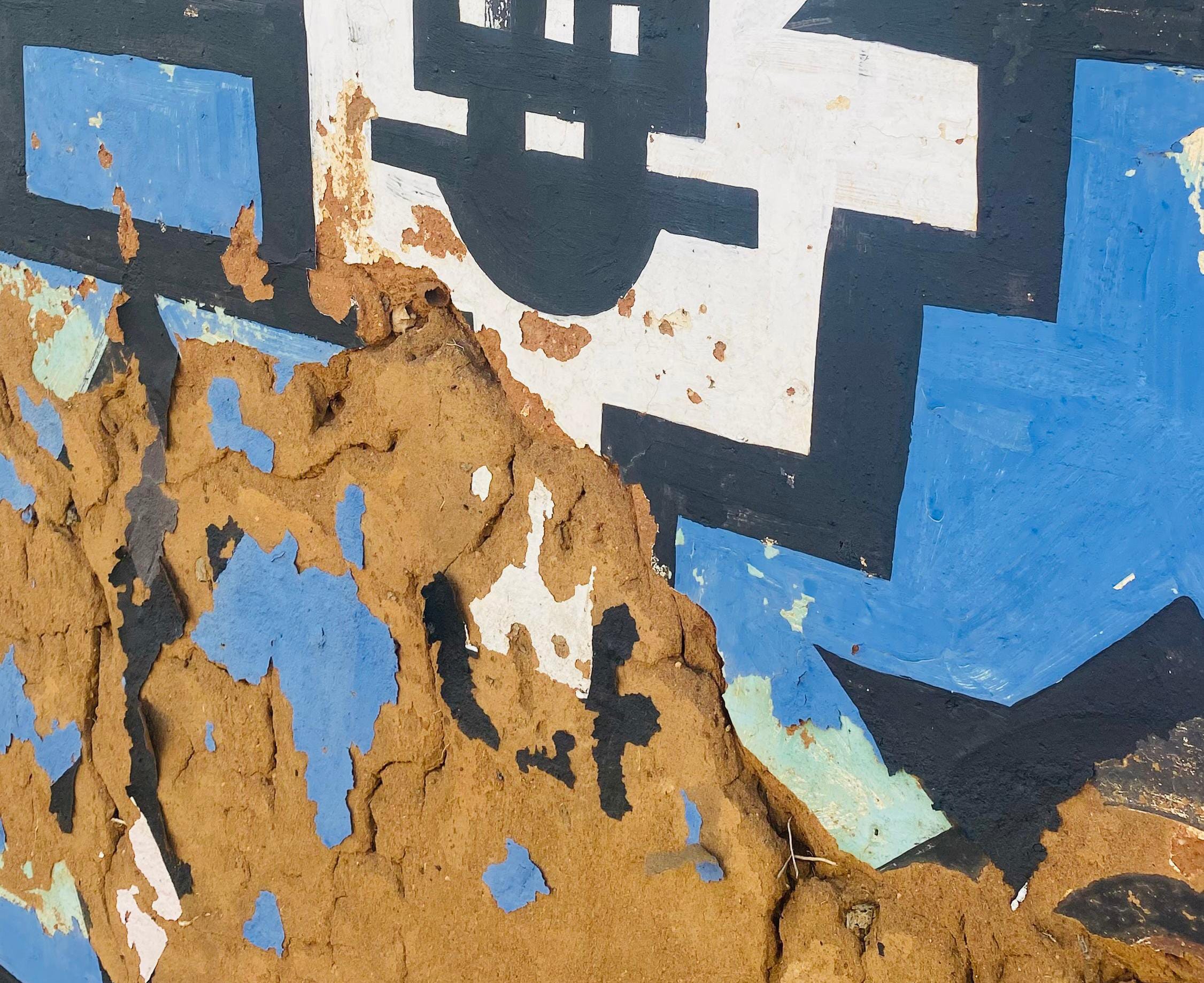 A close-up of a painted wall with geometric black, white, and blue patterns. The chipped paint reveals rough brown mud beneath, giving a weathered look—echoing the enduring spirit celebrated in the Legacy Project of Gogo Mahlangu.