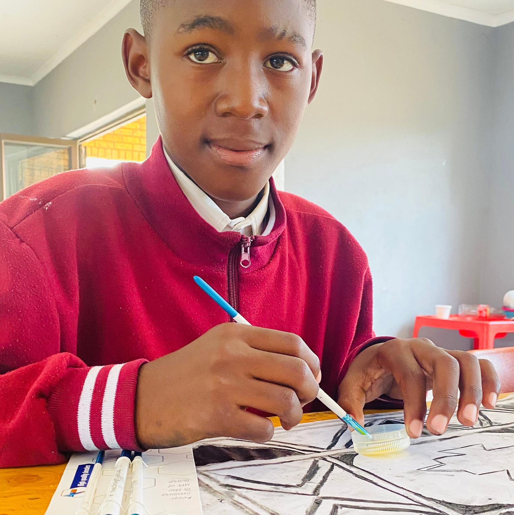 A boy in a red jumper and white collared shirt sits at a table, holding a blue paintbrush and working on a black-and-white geometric drawing. He looks at the camera, as art supplies for the Legacy Project fill the bright indoor space.